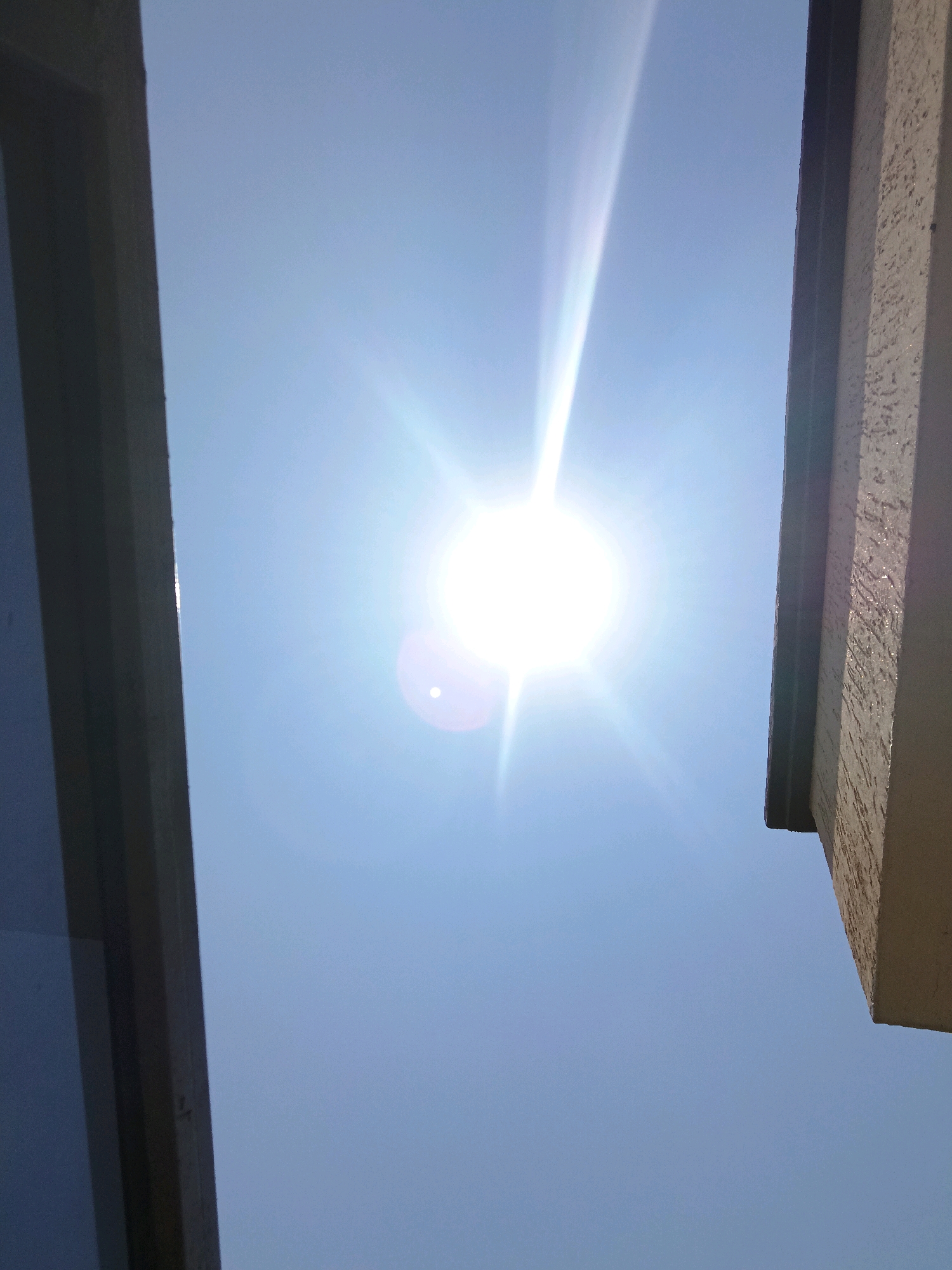 The image shows a bright sun in a clear blue sky, partially obscured by two architectural elements. On the left, a dark, vertical structure, possibly a window frame or a wall, occupies the left third of the frame. On the right, a light-colored, textured vertical structure, likely part of a building's exterior, occupies the right third of the frame. The sun is positioned slightly off-center to the right, emitting strong rays and lens flare. The overall scene suggests an outdoor environment with buildings casting shadows or framing the sky, typical of an urban or suburban setting in Gitaramuka, Burundi. No people or specific actions are visible.