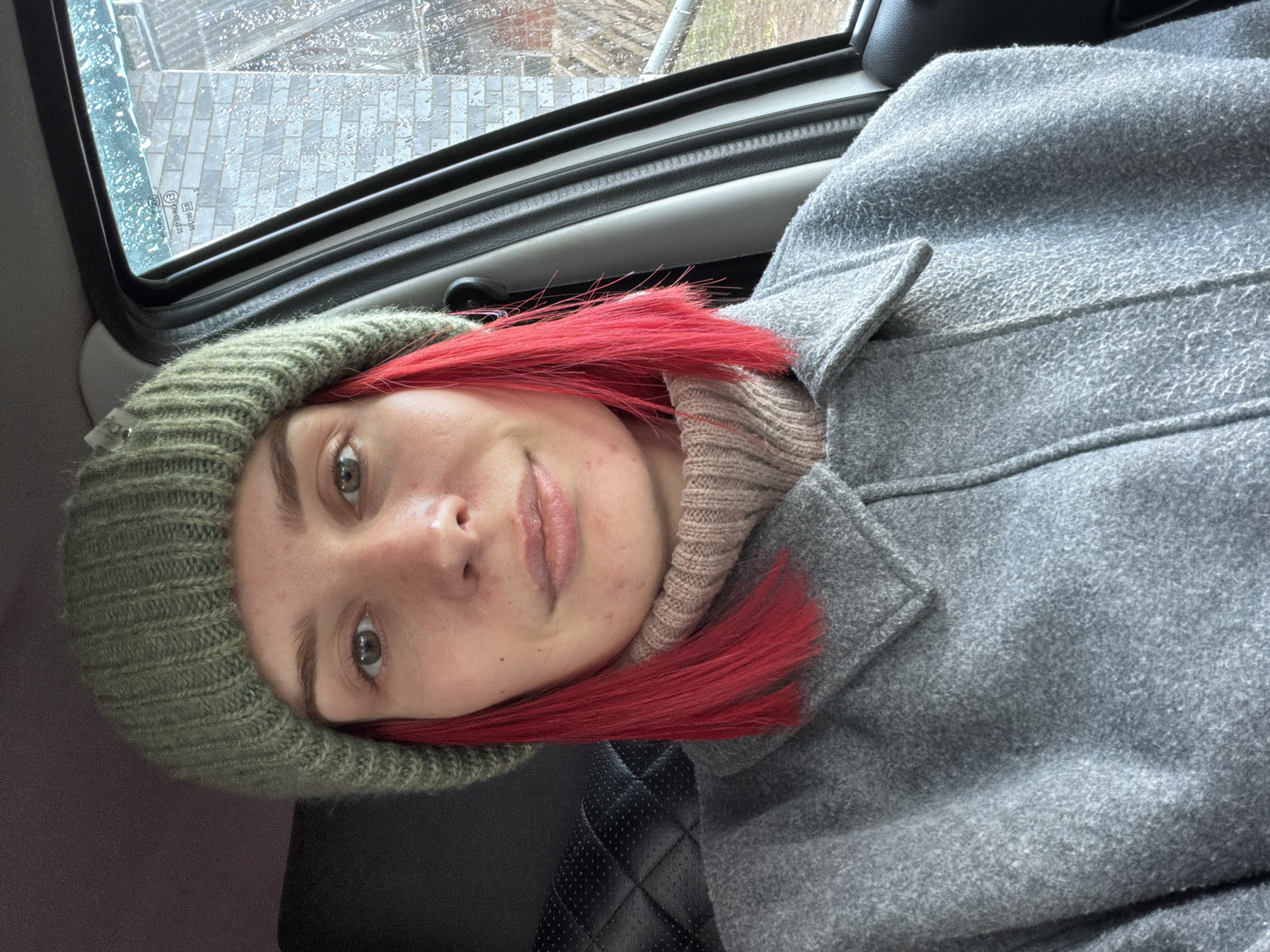 A young individual with bright red hair, light eyes, and a subtle, closed-mouth smile is depicted in a selfie-style photograph taken inside a vehicle in Chaikovsky, Russia. The person is wearing a ribbed olive green knit beanie, a light beige ribbed turtleneck or sweater, and a grey wool-blend coat with a structured collar.
The setting appears to be the interior of a car or bus, with a black quilted leather seat or headrest visible behind the subject, along with grey plastic interior trim and black rubber window seals. Through the vehicle's window, numerous raindrops are visible, indicating rainy or recently rainy weather. The exterior scene shows a wet, grey paved surface, likely interlocking pavers or cobblestones, suggesting an urban or developed environment.
The lighting is soft and diffused, consistent with an overcast daytime sky. Regulatory text and symbols are visible on the car window, including partially legible strings like "E1 00 3075" and "43R-004323". The person looks directly at the camera with a calm and gentle expression.