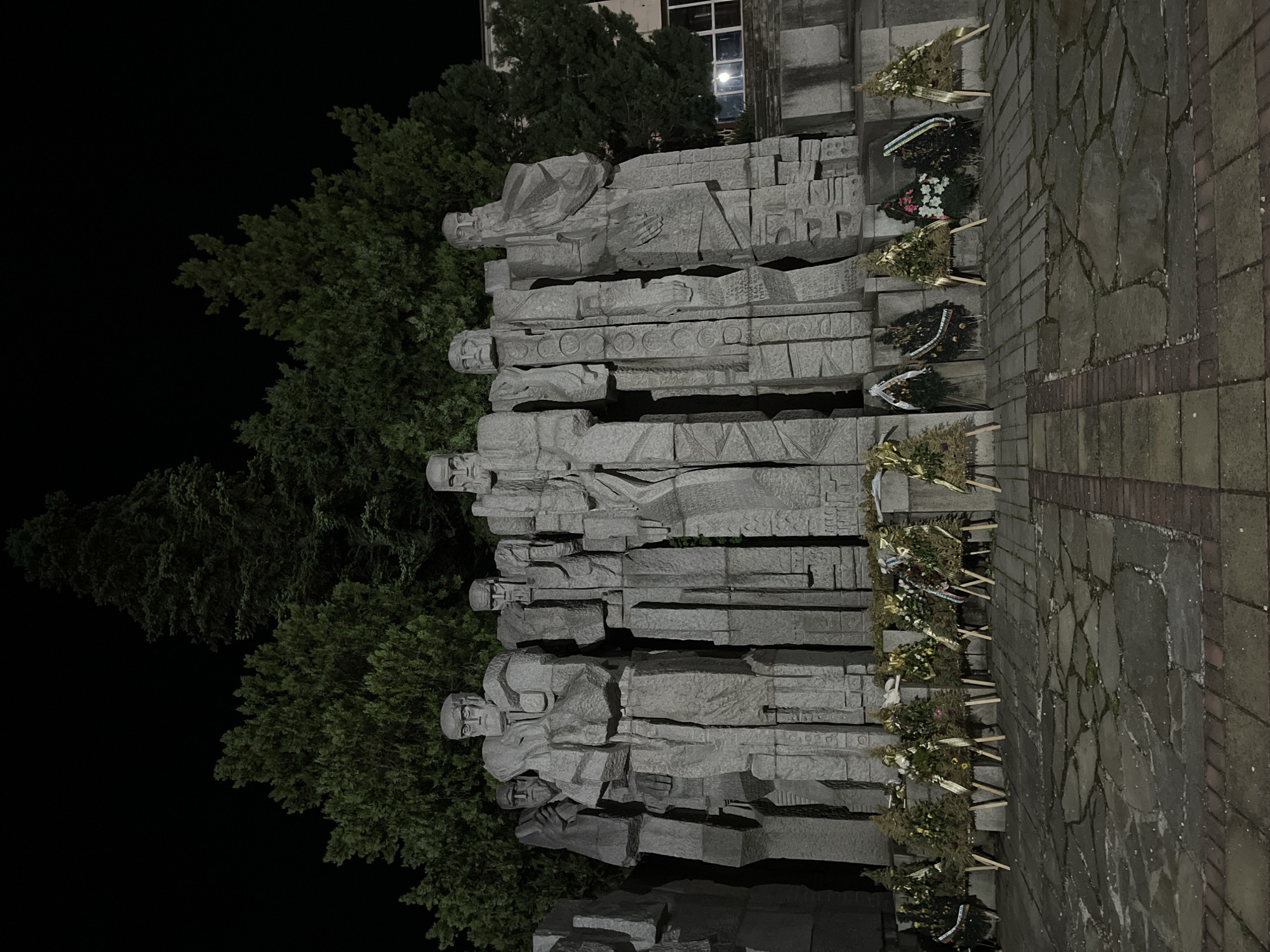This nighttime, low-angle photograph captures a prominent stone monument in Veliki Preslav, Bulgaria, featuring a row of at least seven stylized, blocky male figures. The monument, rendered in grey stone with an angular, Brutalist aesthetic, depicts the figures standing upright with solemn expressions and indistinct, flowing attire, illuminated by artificial light.
In the immediate foreground, arranged along the base of the monument, are numerous commemorative wreaths. These include both triangular wreaths, primarily composed of evergreen branches and decorated with ribbons (some golden, others dark or featuring national flag colors), and circular wreaths. Each wreath rests on a small, light-colored wooden stand. The ground beneath is paved with irregular dark stone slabs and bordered by brickwork, showing signs of dampness.
Behind the monument, the background is dominated by a dense array of tall evergreen trees, appearing dark against the black night sky. On the far right, partially visible, is a building with illuminated windows, suggesting an urban or semi-urban context. The scene conveys a sense of solemn commemoration.