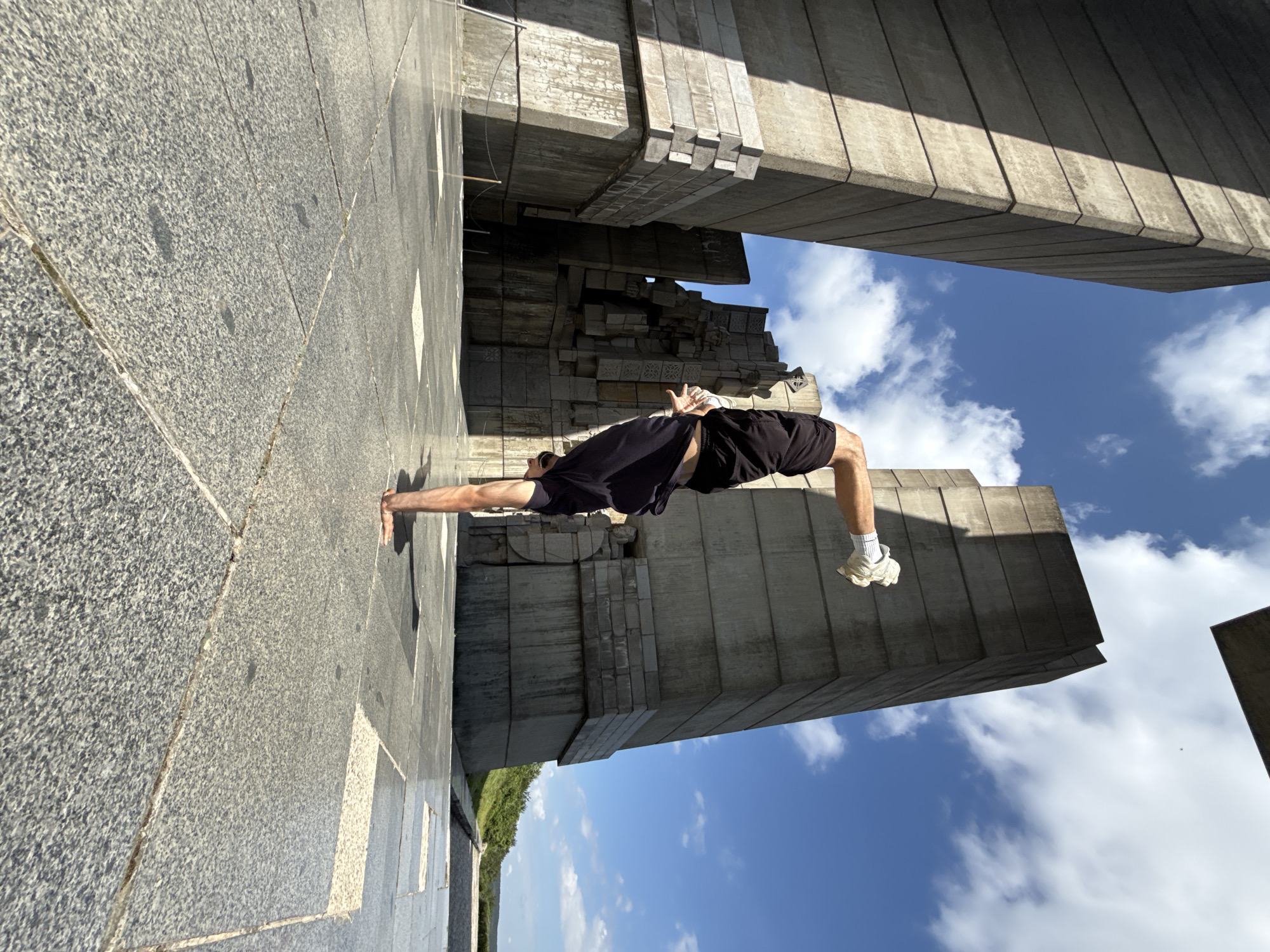 A person is performing a handstand on a paved plaza in front of the Monument to the Declared Bulgarian Communist Party, located in Shumen, Bulgaria. The monument, made of concrete and stone, features imposing, angular structures and is situated under a bright blue sky with scattered white clouds. The person is wearing black shorts and a dark t-shirt, with white gloves on their hands. The plaza floor is made of large, grey granite tiles. The perspective is low, looking up towards the monument and the person.