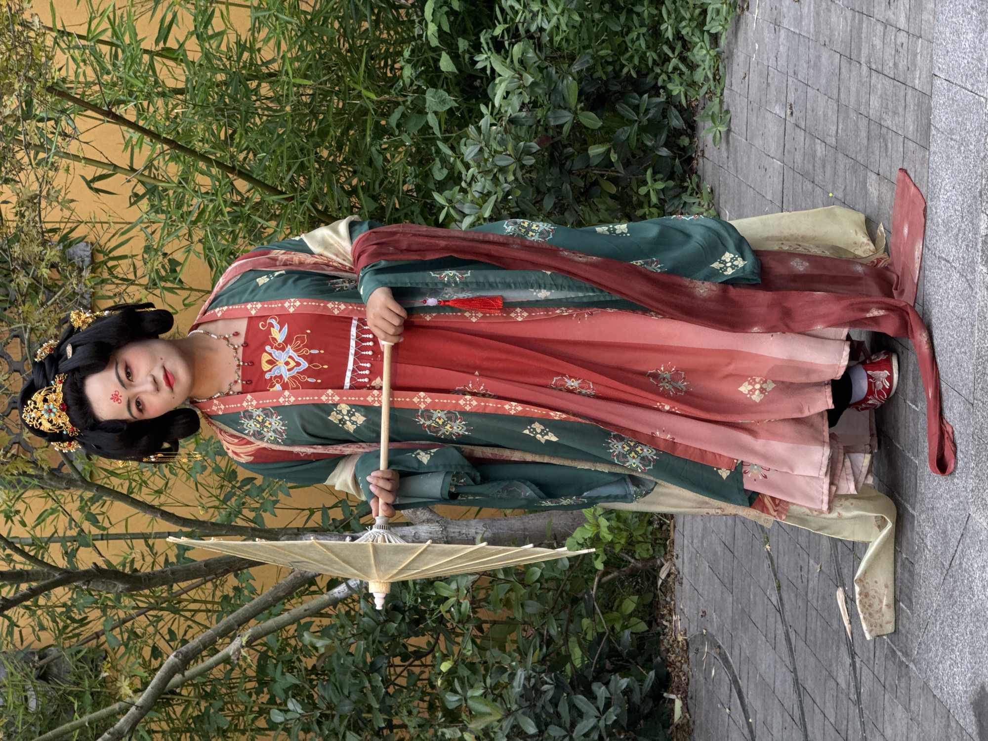 A full-body, upright portrait shows a person in traditional Chinese Hanfu attire, standing in an outdoor setting in Yuzhong District, China. The individual looks towards the viewer with a pleasant, slightly smiling expression. They are adorned in a layered Hanfu dress featuring rich dark green, deep red, and cream/gold fabrics, all intricately embroidered with patterns and gold accents. A sheer red sash is draped over their shoulders.
Their hair is styled in a traditional updo, decorated with ornate gold hair ornaments. Distinctive makeup includes a red forehead mark, defined eyes, and red lips. A red and cream beaded necklace completes the look.
The person gently holds a light cream-colored paper parasol with a bamboo handle in their right hand. The background consists of lush green foliage, including bamboo and bushes, set against a light yellow wall. They stand on a grey flagstone path. The scene is illuminated by soft, ambient daylight. There is no visible text.