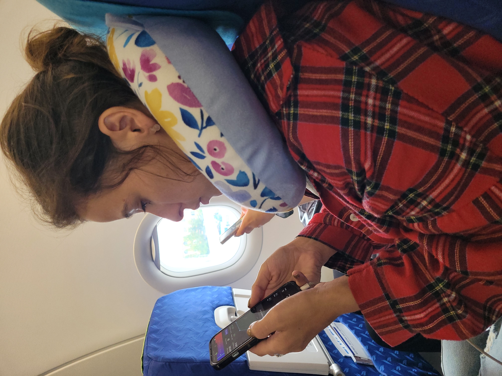 A woman with brown hair pulled into a bun is sitting on an airplane, looking at her phone. She is wearing a red and black plaid shirt and a floral neck pillow. A second person is partially visible in the background, also looking at a phone. The airplane interior includes blue seats and a window showing an outdoor scene. The scene is not in Busan, South Korea, as there are no identifying features of that location.