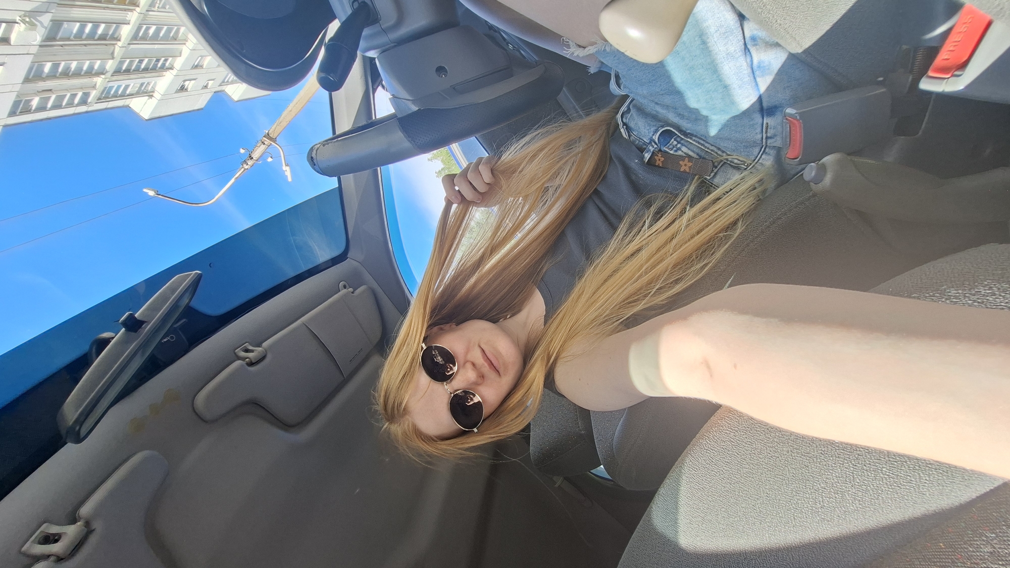 The media file captures a selfie taken inside a vehicle, likely a car, by a young woman with long, blonde hair. She is wearing a gray t-shirt and blue denim shorts, with a belt that has a star-shaped buckle. She is also wearing round, dark sunglasses. Her hair is spread out around her face and shoulders, and she is holding a portion of it with her left hand. The scene is illuminated by natural sunlight, suggesting it is daytime.
The interior of the vehicle is visible, including the dashboard, steering wheel, a rearview mirror, and a sun visor. The windshield shows a clear blue sky with some light rays, and a building with multiple windows and a street lamp are reflected or visible through it. The image is oriented sideways, making the interior of the car appear upside down. The overall mood appears casual and relaxed, possibly indicating a road trip or a moment of downtime. The location context provided indicates Chaikovsky, Russia. No discernible text is visible in the image.