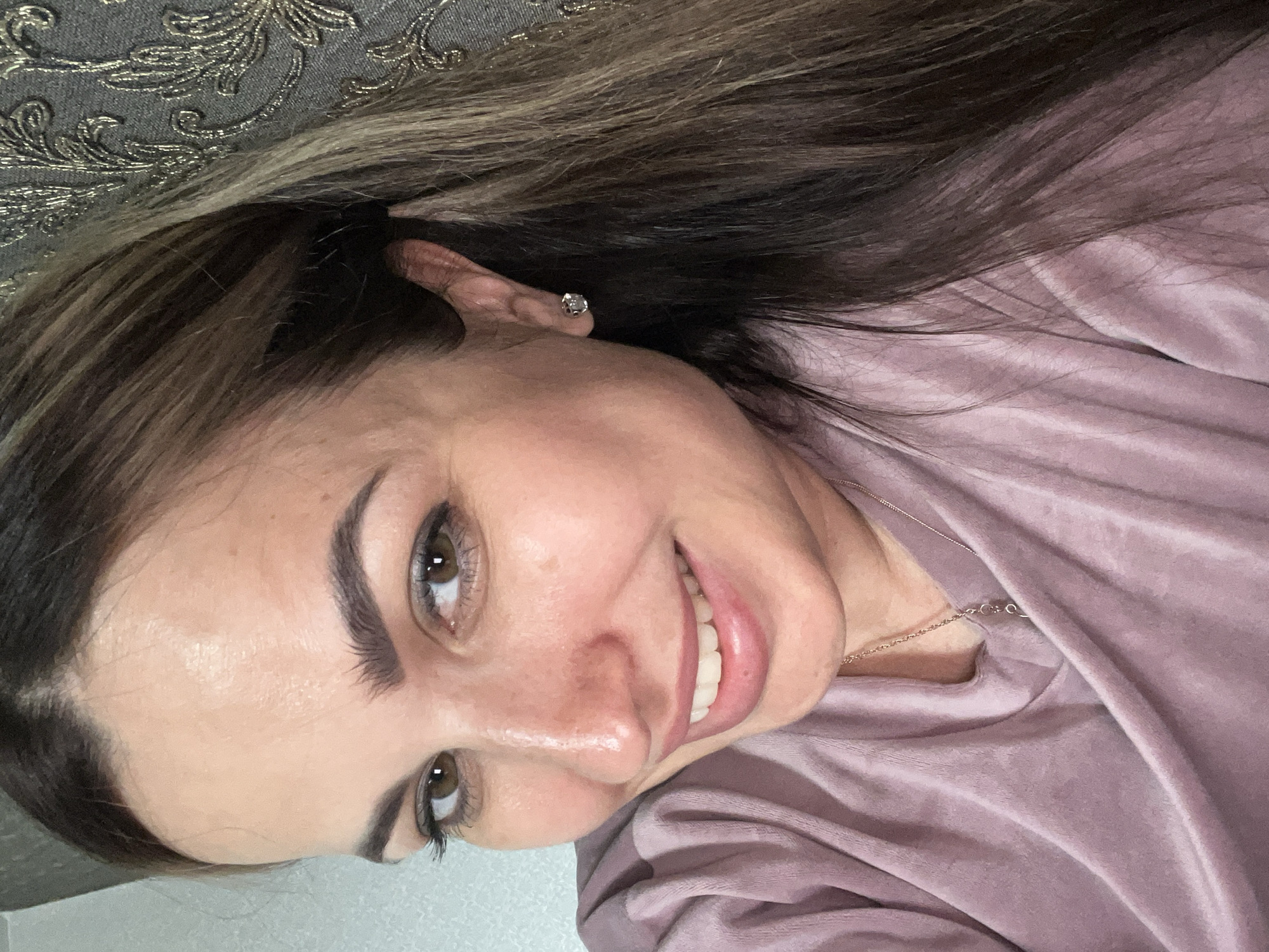 A woman is captured in a close-up, head-and-shoulders selfie. She has brown hair with lighter streaks, pulled back and falling over her right shoulder. Her eyes are a light brown and she is smiling, revealing her teeth. She is wearing a light purple velour top and a silver necklace with a pendant. A small, diamond stud earring is visible in her left ear. The background is a textured, patterned wallpaper in shades of grey and gold. The image appears to have been taken indoors, in a well-lit environment.