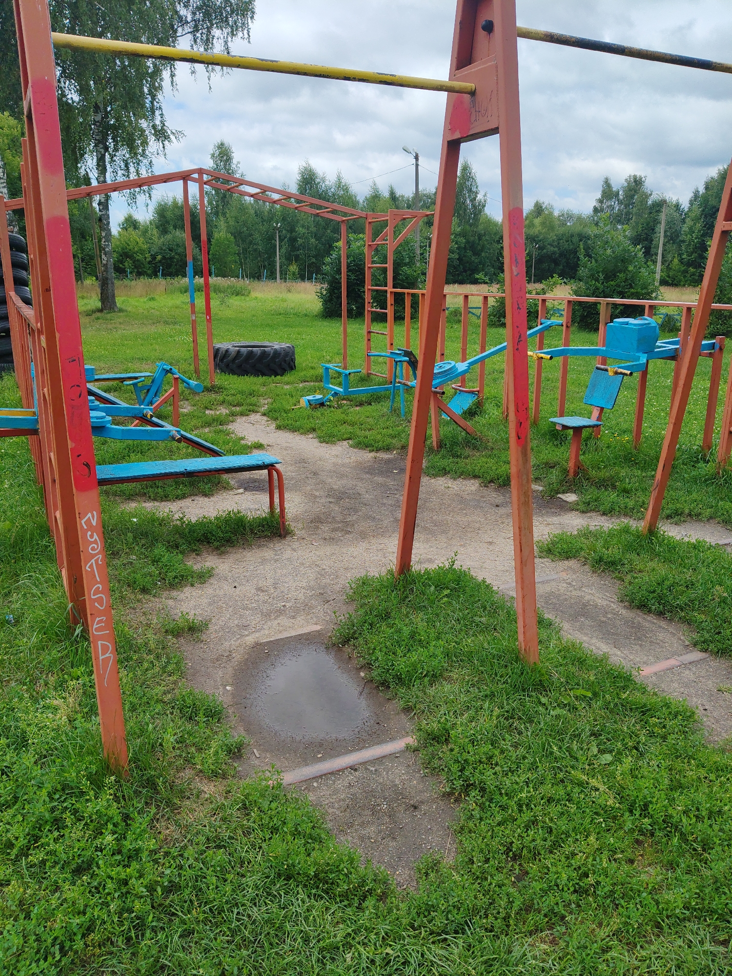 The image shows an outdoor fitness equipment area in Drezna, Russia. The equipment is made of red and blue metal frames, with some yellow bars. There are monkey bars, a bench press, a seesaw, and other exercise machines. The area is surrounded by grass and trees. The ground is a mixture of dirt paths and concrete. There is a puddle of water on one of the paths. The sky is cloudy. No people are visible in the image.