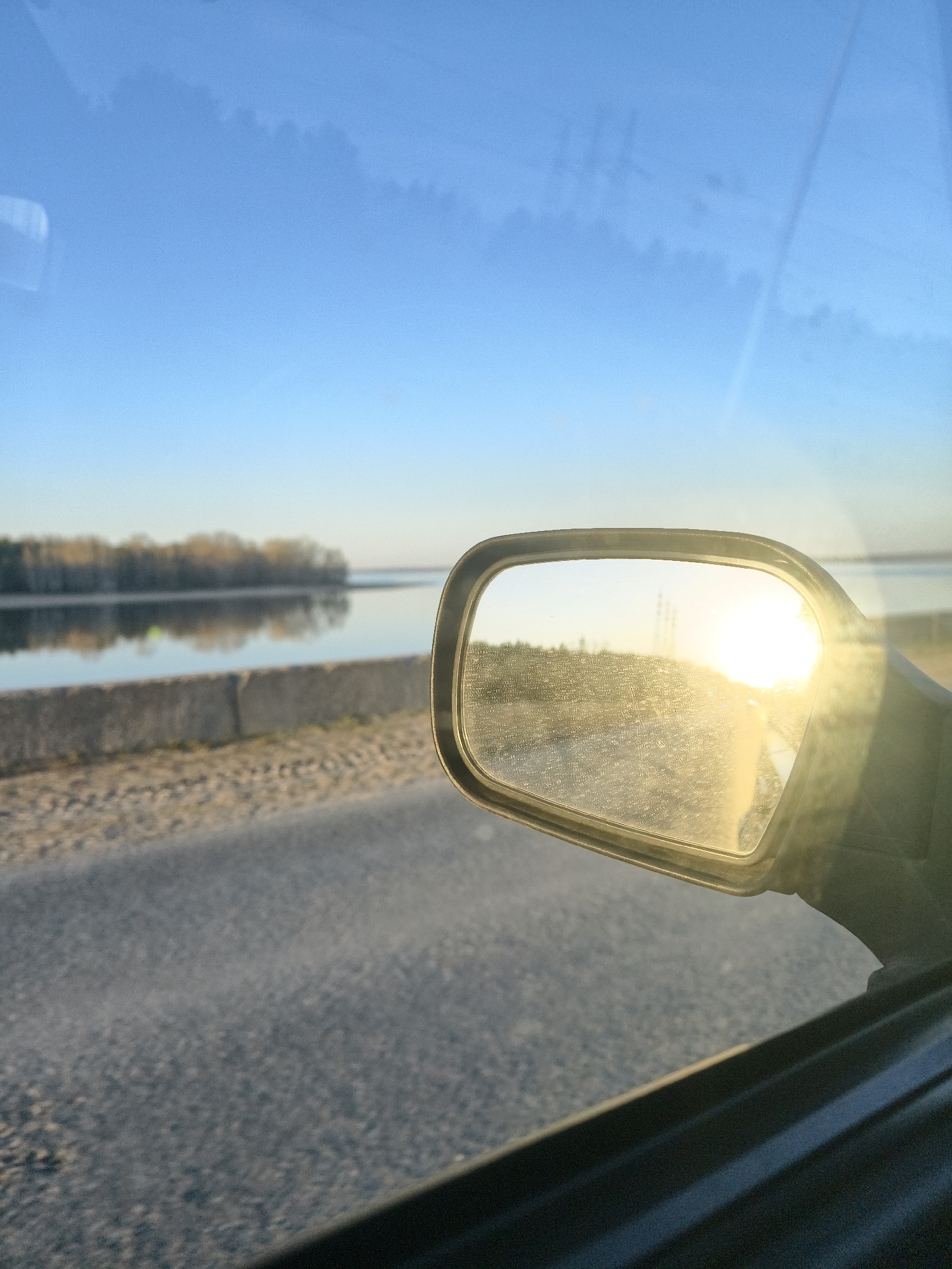 The image depicts a view from inside a vehicle, possibly a car. The asphalt road and a concrete barrier are visible through the side window, with a body of water and trees on the other side of the barrier. The side mirror reflects the sun and land, possibly a road or field. The sky is blue, with some wispy clouds. The location is identified as Noviy, Russia.
