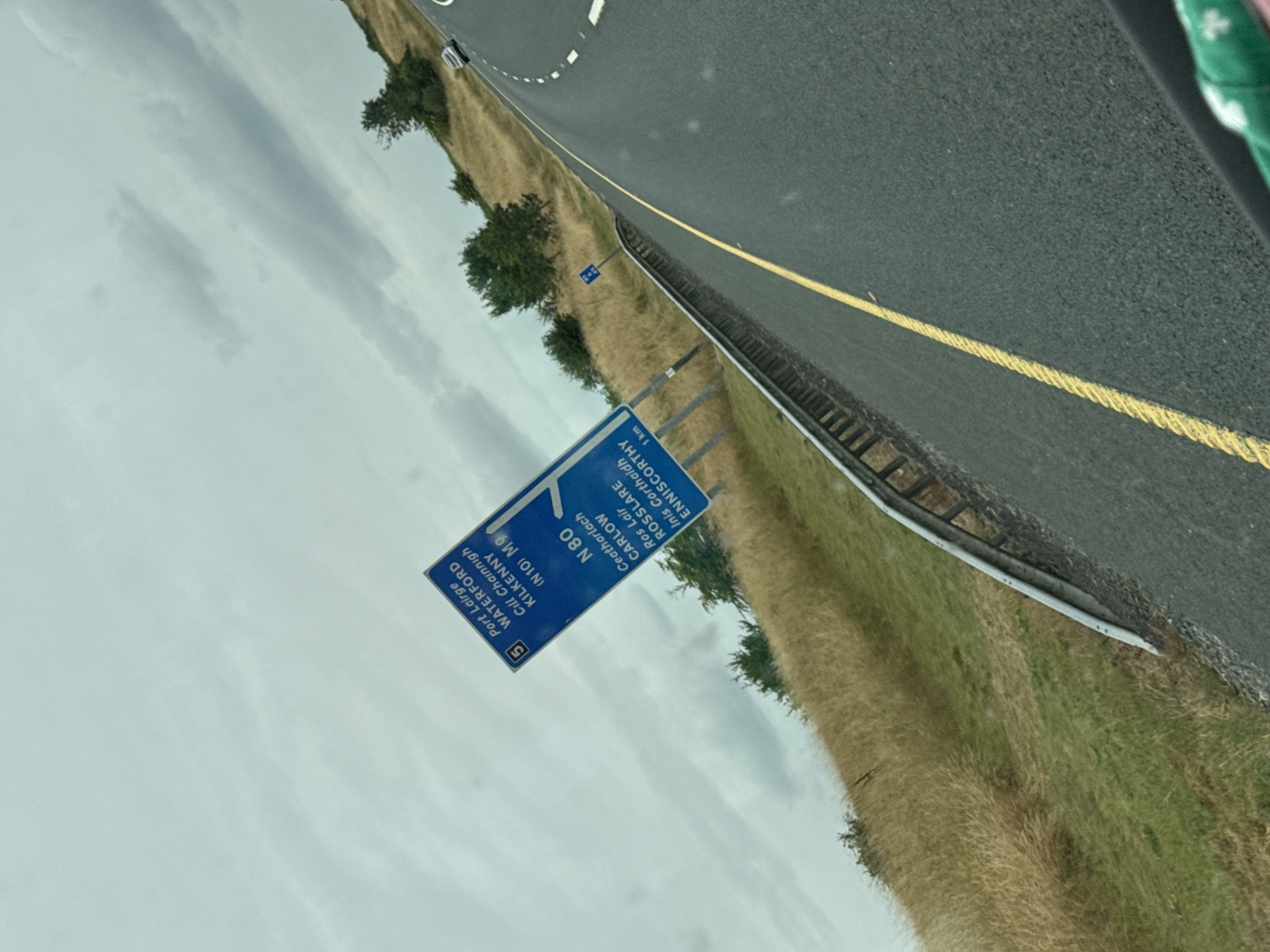 The image shows a road sign on the side of a road in the city of Moyle, Ireland. The sign is blue with white lettering and indicates directions to various towns. The road is asphalt with a yellow line down the middle. The surrounding area is grassy with some trees and a cloudy sky. The image is taken from a car.