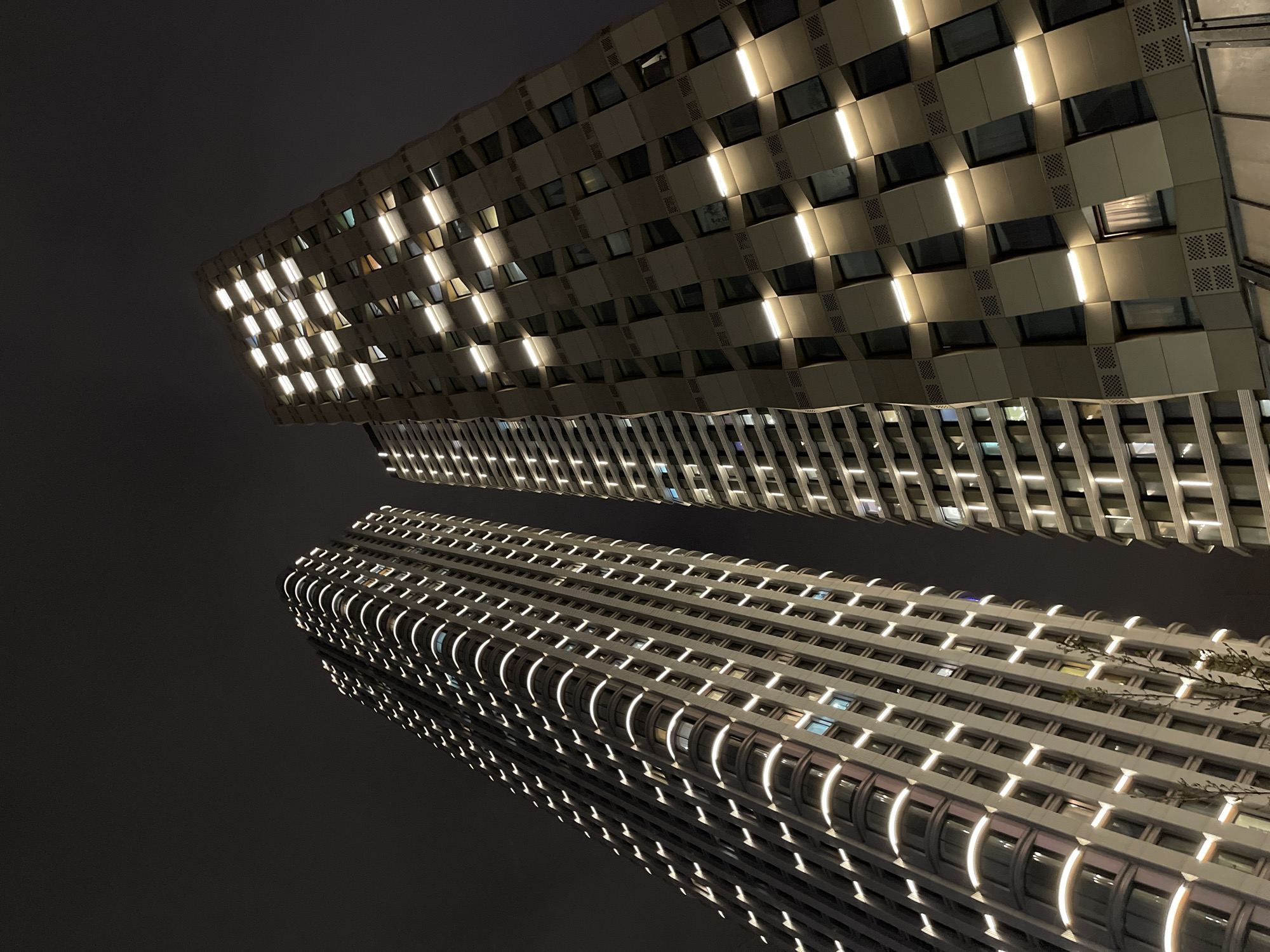The image depicts two tall buildings at night. The building on top has a geometric facade of interlocking squares and rectangles with intermittent vertical lighting. The lower building features a more traditional rectangular structure with curved balconies and linear lighting. The overall composition is an upward view, showcasing the height and architectural details of the buildings. It is nighttime, as the buildings are illuminated against a dark sky. While the information provided states the location is Khimki, Russia, this detail cannot be determined from the image alone.