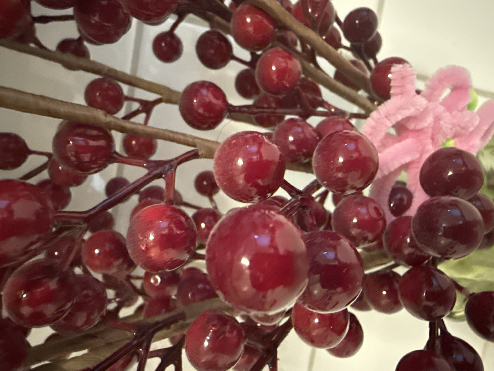A close-up, high-angle shot shows a cluster of dark red, artificial berries on thin, brown stems. The berries are round and have a glossy finish, reflecting light. Some are in sharp focus, while others are blurred. In the lower right, a portion of a pink, fuzzy object, possibly a craft flower, is visible. The background is a pale, out-of-focus white. There are no people or animals in the image. The scene is devoid of any specific environmental context that would indicate a location in Ridabu, Norway, or any other city.