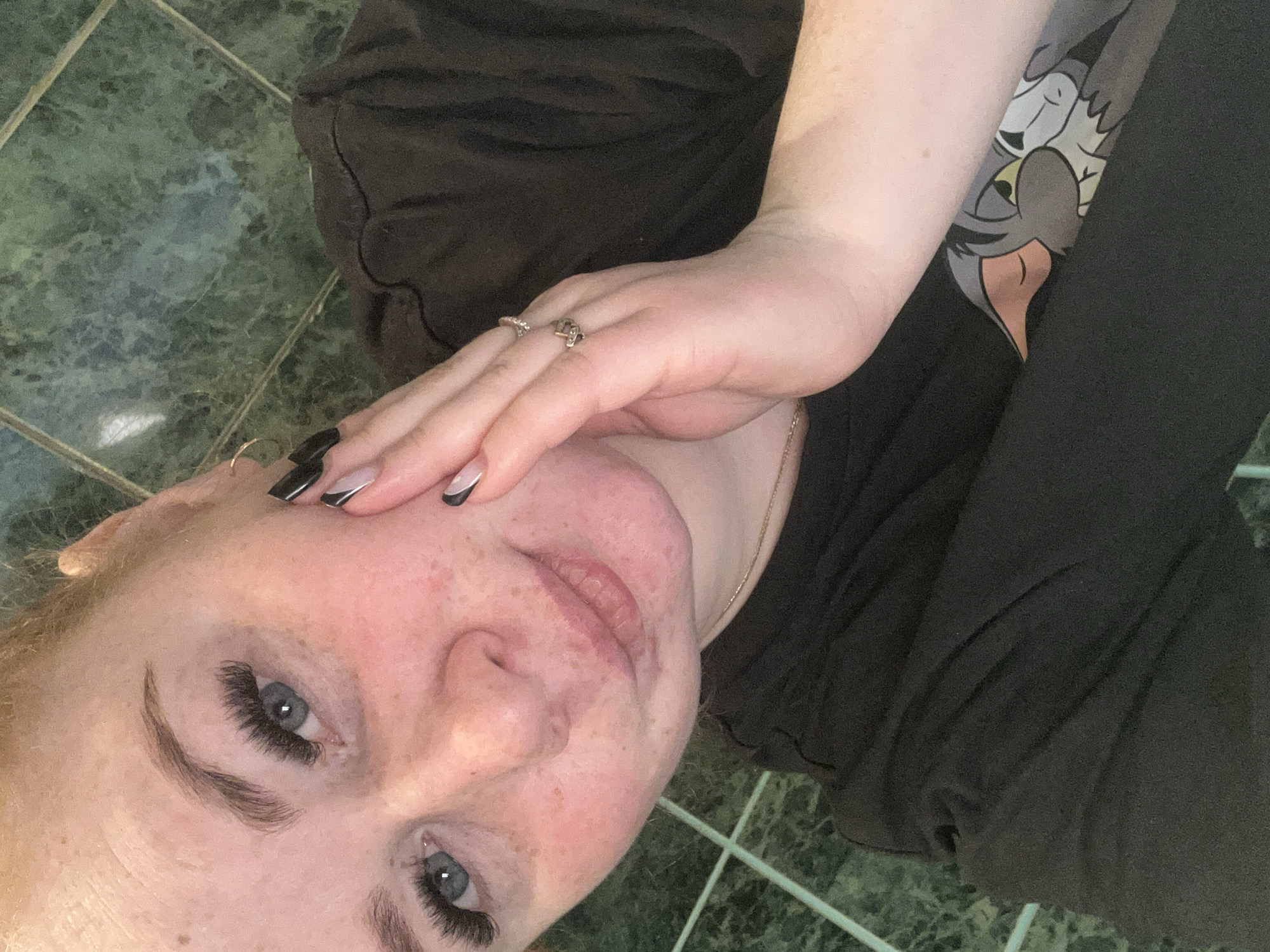 A close-up selfie of a fair-skinned person with freckles and light eyes, oriented at an angle. The person is wearing false eyelashes and has black and white nail polish. Rings are visible on their fingers. They are wearing a dark-colored shirt with a cartoon graphic on the sleeve. The background appears to be a green tile floor with white grout lines. There is no indication in the image that it was taken in Chaikovsky, Russia.