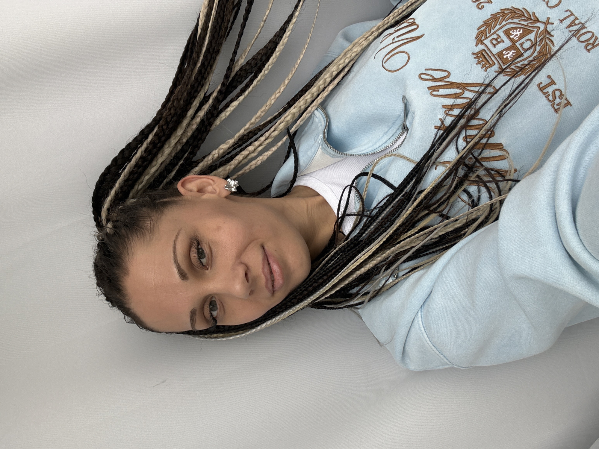 A woman is depicted in a medium close-up, oriented on her side with her head turned towards the camera. She has warm-toned skin and long, extensive hair styled in numerous small braids, featuring a mix of black, dark brown, and light blonde or beige colors. Her gaze is direct and calm, accompanied by a subtle, soft smile. She has well-defined eyebrows and appears to wear light eye makeup.
She is dressed in a light blue zippered hoodie, worn over a white top. The hoodie features intricate embroidery in brown thread. Visible details of the embroidery include a crest or emblem with the letters "R" and "C", along with partial text that reads "ROYAL" and "EST". There is also stylized script embroidery that is not fully decipherable.
A small, decorative earring, possibly silver-colored and shaped like a flower or star, adorns her right ear. The background is a plain, light-colored surface, likely a white or light grey fabric, suggesting an indoor setting. The lighting is soft and even, contributing to a relaxed and casual atmosphere. The image's orientation shows the woman's head on the left and her body extending to the right. While the location context is Rusanovo, Russia, no specific visual cues within the image confirm this geographical detail.