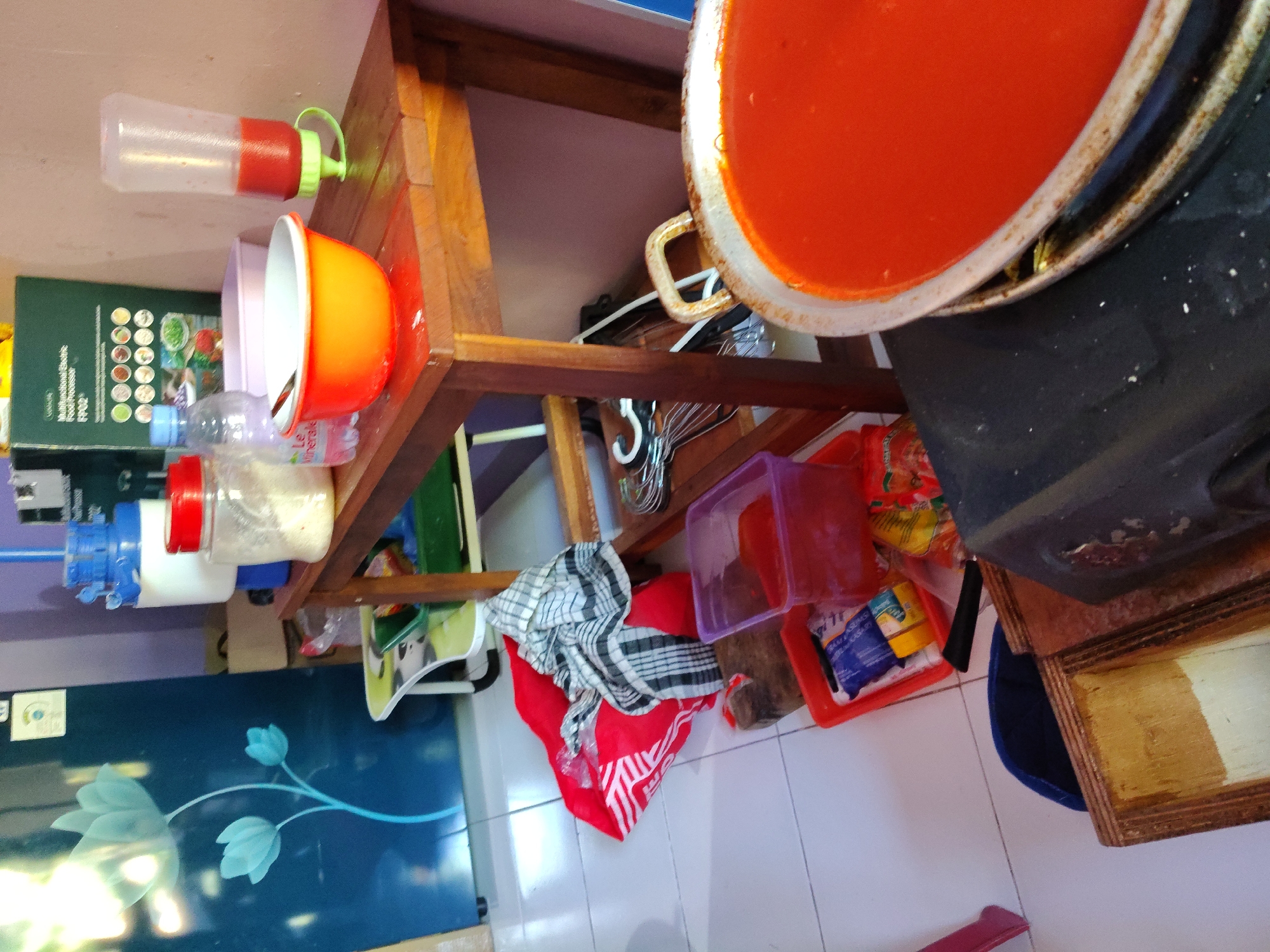 The image depicts a cluttered kitchen counter or utility area. In the foreground, a large pot filled with a bright red liquid, possibly sauce or soup, is placed on a black stove top. To the left of the stove, a wooden box is visible. Behind the stove, a wooden shelf unit holds various items, including a large metal bowl containing a red liquid, a plastic bottle with a red dispenser cap filled with a similar red liquid, a white bowl with a spoon, a clear plastic bottle of water, and a jar with a red lid containing a white granular substance. In the background, a refrigerator with a floral design is partially visible, as well as a blue water dispenser with white accents. Various containers, bags, and cloths are scattered on the floor and counter. The scene suggests a busy and possibly domestic environment. The image is shot from a medium-low angle, looking slightly upwards. The overall impression is one of a functional, albeit somewhat disorganized, kitchen space. The presence of Indonesian packaging and branding on some items, as well as the style of the refrigerator and kitchenware, strongly suggests the location is in Indonesia. While no specific landmarks are visible, the everyday items and general ambiance are consistent with a residential kitchen in a city like Surabaya.