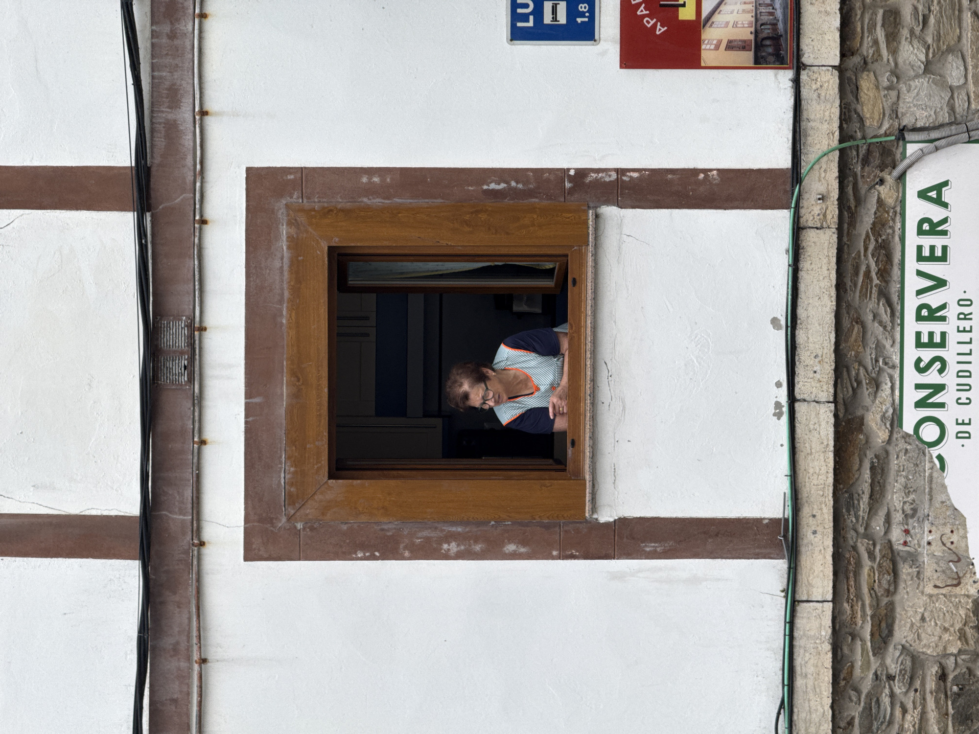 An elderly woman with glasses and short brown hair, wearing an apron over a striped shirt, peers out from an open window of a white building with brown trim. She is leaning on her forearms on the windowsill. To the right of the window, a blue sign with a bed icon and the number "1,8" is visible, as is a red sign with text that appears to be "APA..." and an image of buildings. Below the window, a stone foundation is visible, and further down, a sign reads "CONSERVERA DE CUDILLERO". The scene suggests an urban setting in Cudillero, Spain, possibly a residential or commercial area.