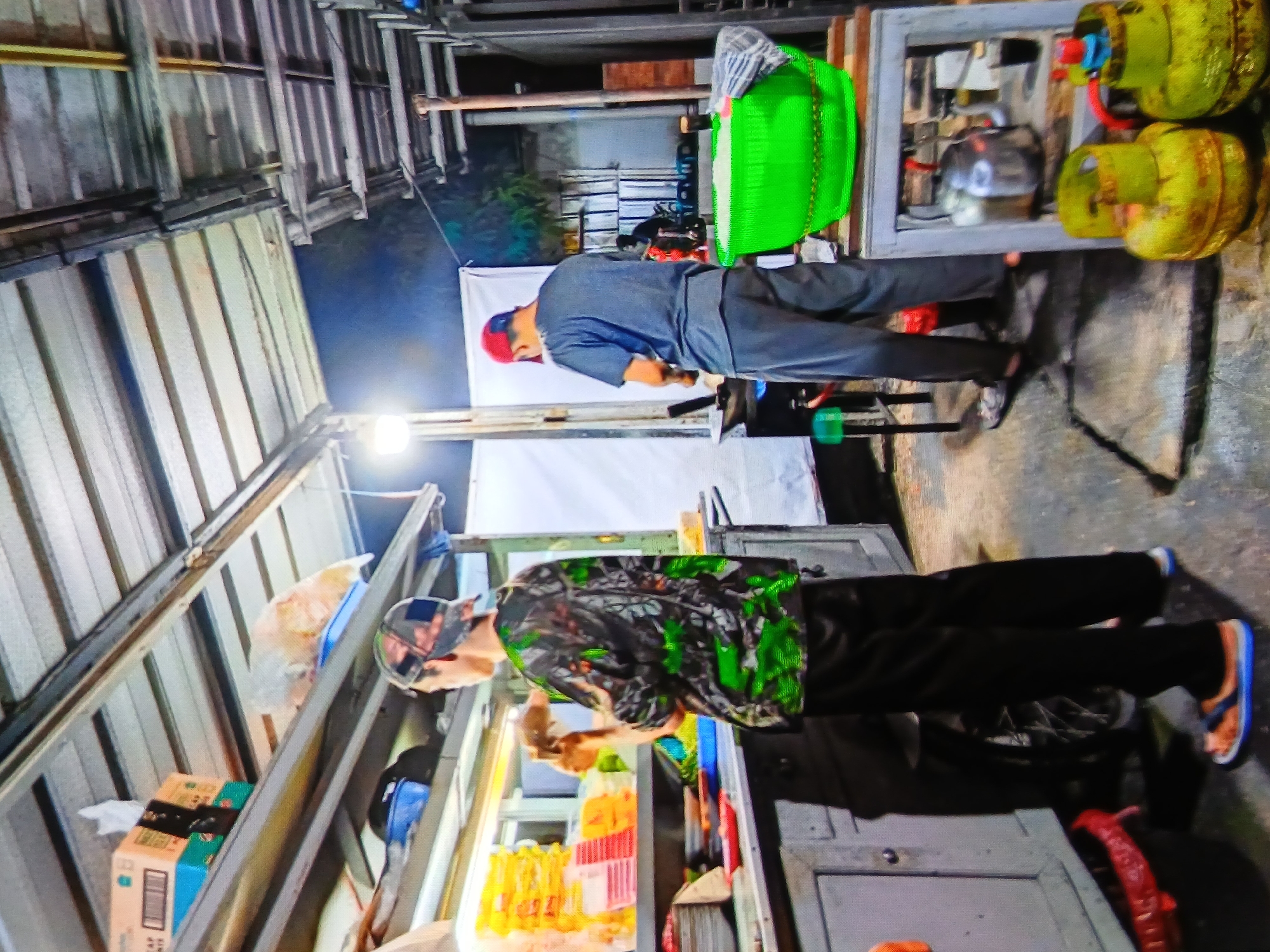 The image depicts an outdoor food stall at night in Setia Bakti, Indonesia. Two men are actively engaged in food preparation. In the foreground, a man wearing a camouflage-patterned shirt and black pants is standing behind a food counter, holding a piece of food in his hands. To his right, a man in a grey t-shirt and grey pants, wearing a red cap, is cooking on a stove. The stall is equipped with various food items, cooking utensils, and two large green propane tanks. The background shows the typical urban environment of an Indonesian city at night, with metal roofing and lighting.