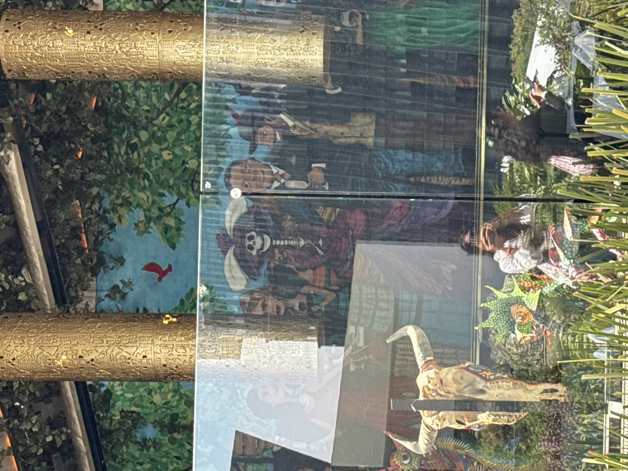 The image shows a window display in Mexico City, Mexico. The display features a combination of art pieces and decorative elements reflecting Mexican culture and history. The glass pane is reflecting the exterior.
Art pieces include a skull with horns, possibly of a goat or ram, suggesting Day of the Dead or other indigenous motifs. There are other artistic representations, possibly portraying people in historical Mexican attire.
The display is framed by a metallic cylinder with patterns, possibly Aztec or Mayan in origin, mounted with light fixtures. A backdrop of greenery is visible, with a red bird appearing to be part of a mural or painting.
The overall scene gives a sense of a blend of traditional Mexican themes and contemporary art.