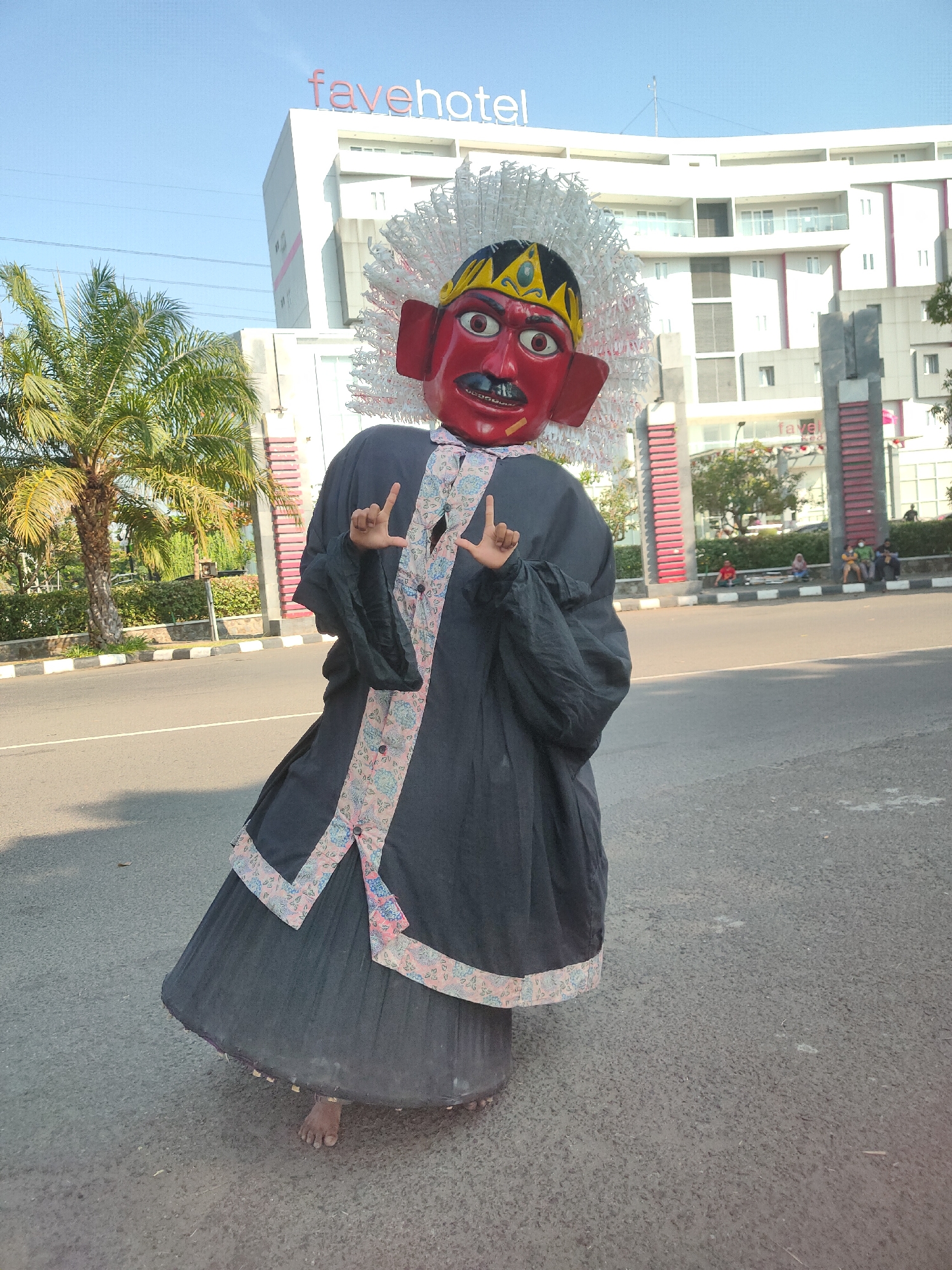 A person in a large, traditional costume stands in the street in front of a fovehotel in Ngasem, Indonesia. The costume is predominantly black with a long, flowing skirt and wide sleeves. It features a red mask with a large headdress of white, feather-like material. The mask has large red eyes, a black mustache, and a yellow and black crown. Decorative trim with a floral pattern in pink, blue, and white runs along the edges of the costume's front opening. The individual's bare feet are visible beneath the hem of the skirt. They are holding their hands up, with fingers extended in a gesture. The street is paved and appears to be a public road. In the background, a white building with the "fovehotel" logo is visible. Palm trees and greenery are also present. Several people are sitting on a curb in the middle distance. The sky is clear and blue.
