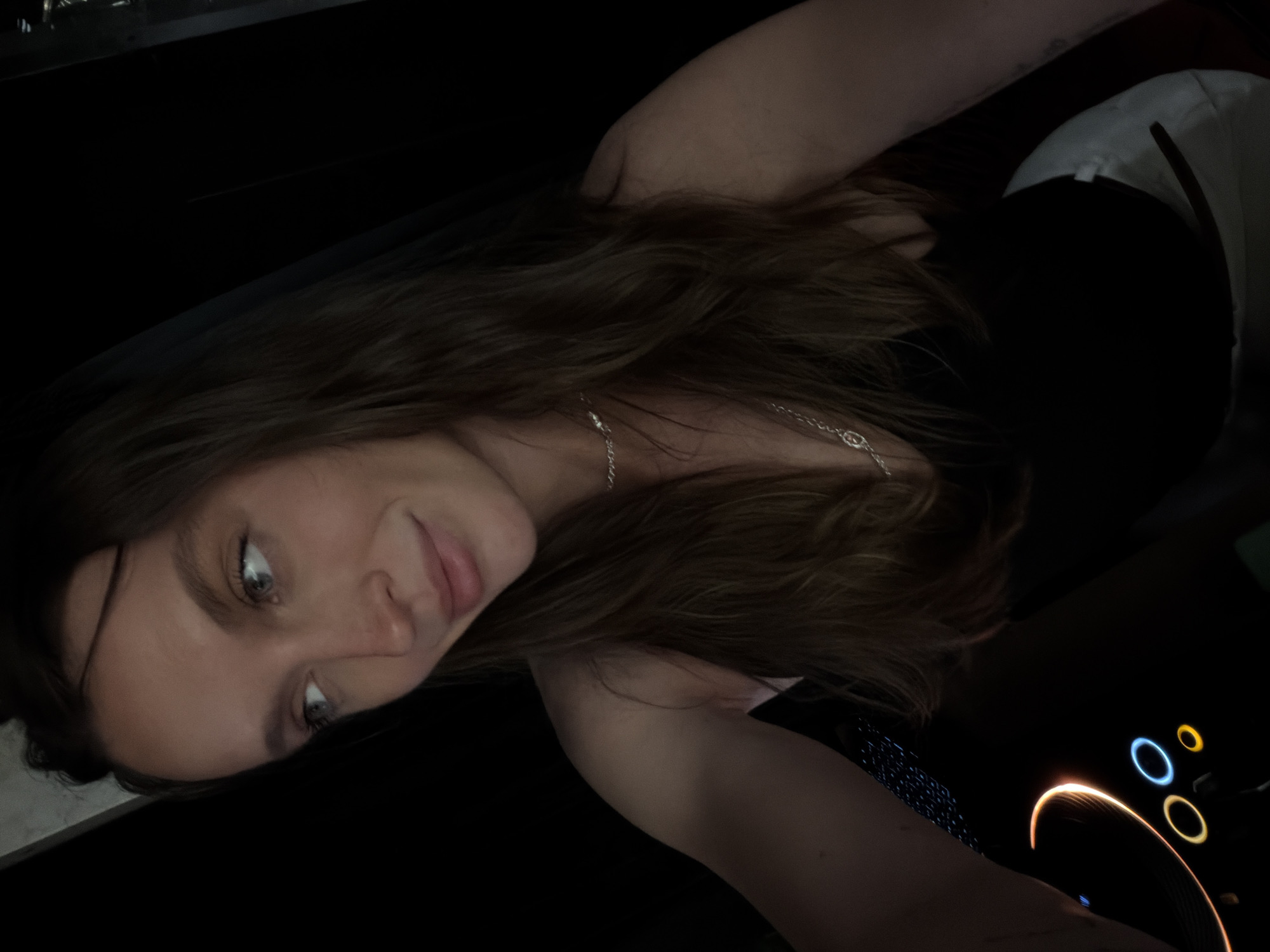 A young woman with brown hair and blue eyes looks at the camera. She is wearing a black top and a silver necklace. To her right is a laptop with a glowing trackpad and illuminated buttons. The background is dark, and the exact location in Budva, Montenegro cannot be determined from the image.