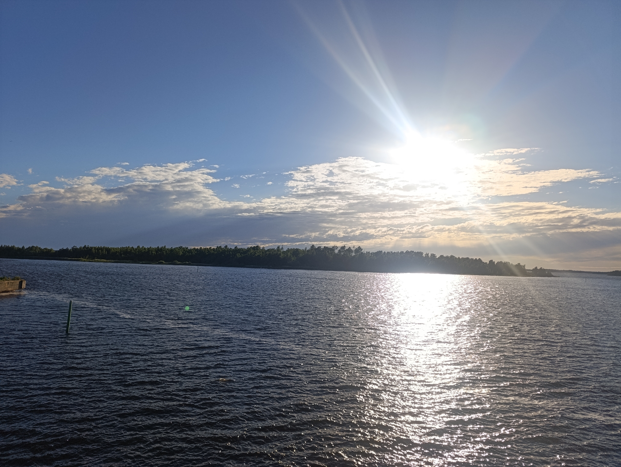 The image depicts a wide expanse of dark, rippling water, likely a bay or large lake, under a bright, sunlit sky. The sun, positioned in the upper right quadrant, emits strong rays that create a shimmering reflection on the water's surface. A line of trees forms a dark silhouette along the distant horizon, marking the edge of land. The sky is a gradient of blue, with scattered white clouds, some of which are illuminated by the sun, creating a dramatic contrast. A green post is visible in the water on the left side of the image, and a small concrete structure is also present on the far left edge. The overall impression is one of a serene, natural landscape at a time of day when the sun is low, casting dramatic light and shadow.
This scene is the Alholmen bay area in Jakobstad, Finland, a location known for its waterfront views and natural beauty.