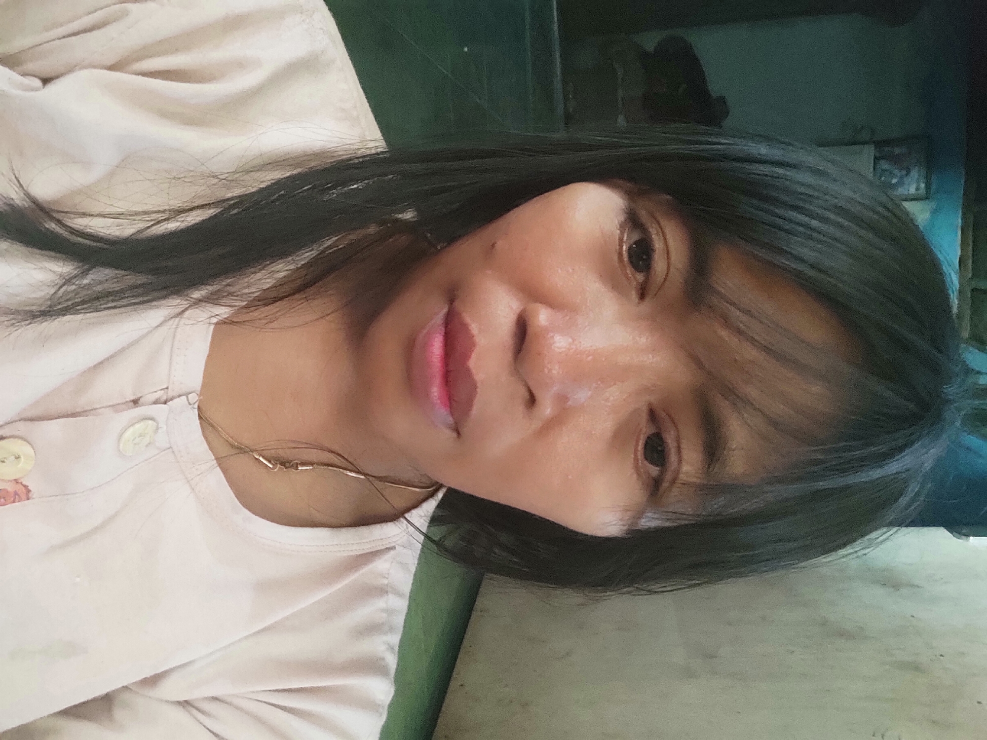 A selfie taken from a frontal perspective shows a woman in Sakra, Indonesia. The woman has dark hair that falls around her face, and she is wearing a light-colored shirt with buttons. She appears to be indoors, with a textured wall behind her on the right and a darker, possibly cluttered area on the left. The lighting suggests it might be daytime, but the indoor setting limits definitive conclusions about the outdoor environment.