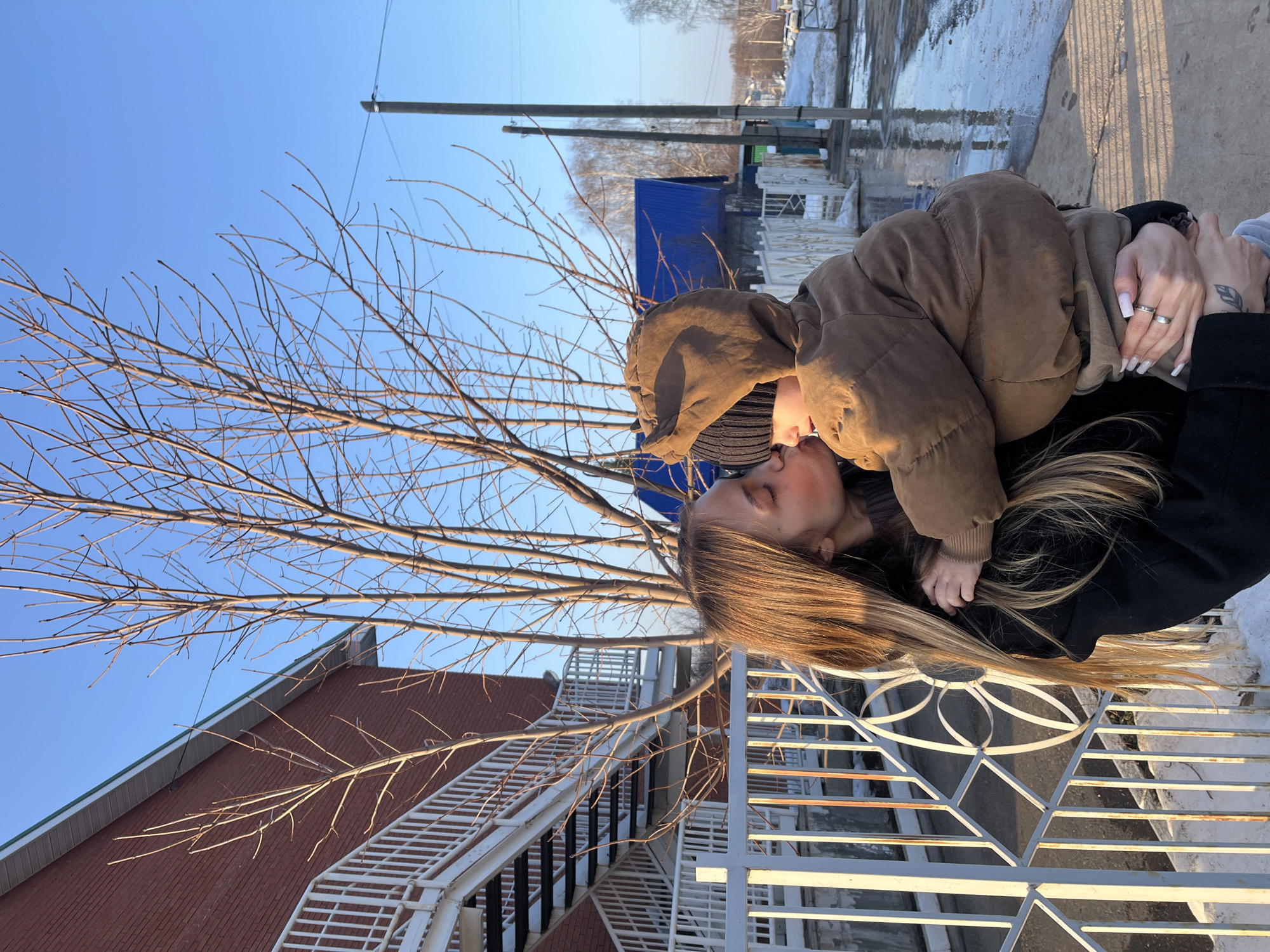 An outdoor daytime image taken in the city of Фоки, Russia, shows an adult female holding a young child. The female, positioned on the left, has long, light brown hair illuminated by sunlight and wears a dark coat. She is seen with her head turned, appearing to kiss the child's right cheek. A silver-toned ring is visible on her left ring finger, and a leaf-shaped tattoo is present on her left forearm. The child, held in her arms, wears a brown hooded puffer jacket and a dark knit beanie, with their face largely obscured by the adult's head and hood.
In the foreground, a white metal fence with decorative circular elements runs horizontally. Behind the subjects, to the left, is a red brick multi-story building featuring a white trim along its roofline and an external white metal staircase with multiple landings and railings. Several deciduous trees with bare branches stand in the mid-ground. To the right, a wooden utility pole with wires is visible, with a house featuring a bright blue corrugated metal roof further in the background. The ground appears wet with puddles, patches of melting snow or ice, and visible tire tracks and footprints. The sky is clear and bright blue, indicating sunny weather.