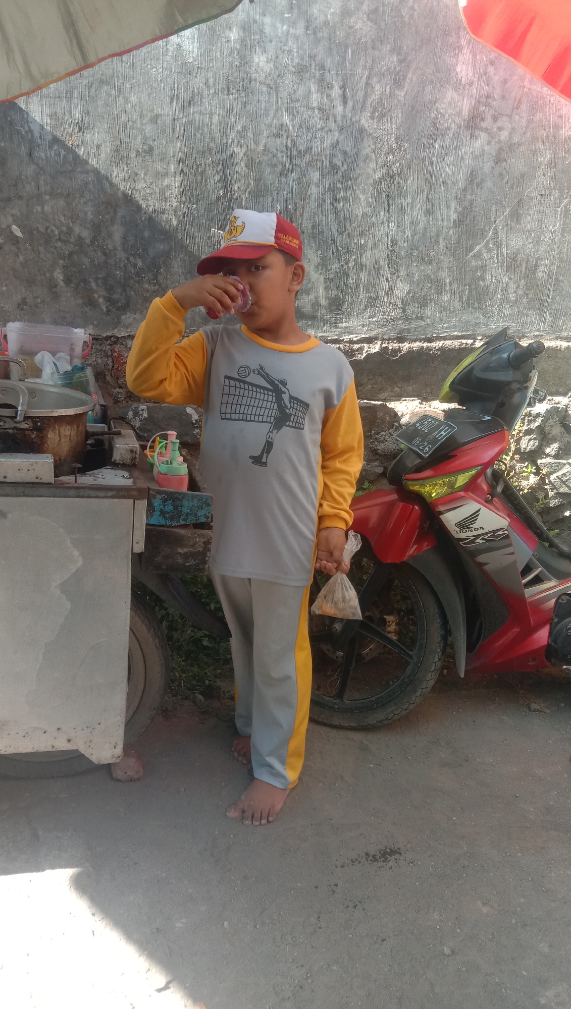 A young boy, likely under ten years old, stands barefoot on a dirt and concrete surface in front of a grey textured wall. He is wearing a long-sleeved grey shirt with yellow sleeves, featuring a graphic of a volleyball player. He also wears grey pants with a yellow stripe down the side and a red and white baseball cap. The boy is holding a clear plastic bag containing food in his right hand and drinking from a small plastic cup with his left. To his left, a metal food cart with various containers and cooking implements is visible. To his right, a red and black motorcycle is parked. The scene suggests an outdoor, possibly roadside, setting in Karanganyar, Indonesia, indicated by the style of the food cart and the presence of the motorcycle, common elements in Indonesian street scenes. The lighting indicates it is daytime, with shadows cast by an overhead structure, possibly an awning.