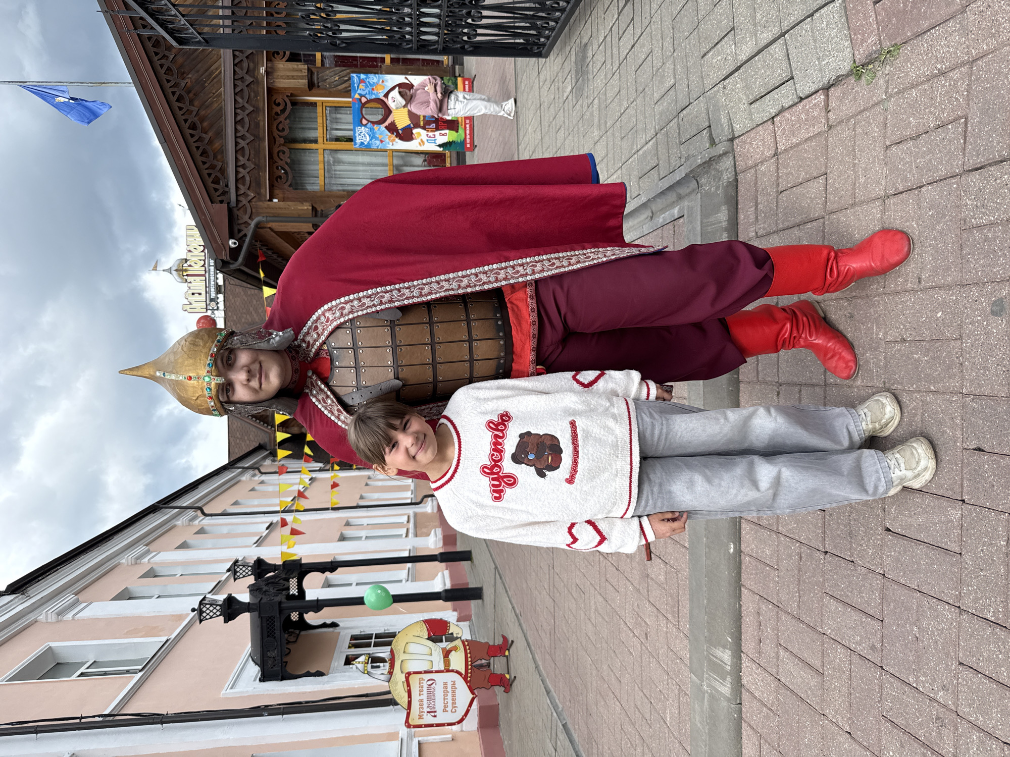 The image shows a full shot of two individuals and part of a streetscape. A girl stands in front, wearing a white sweater with a bear design and grey trousers, with white sneakers. Behind her is a person in a traditional Russian costume, wearing a helmet, armor, a red cloak, and red boots.
The setting appears to be outdoors on a brick-paved walkway. Buildings are visible in the background, including one with decorative wooden architecture and colorful triangular flags or banners hanging above the street. There's a sign with a caricature of a knight with text written in Russian. There's also a sign with Russian writing on it.
Based on the limited details, it is difficult to confirm the specific location without additional clues, but the image strongly suggests it is Rusanovo, Russia.