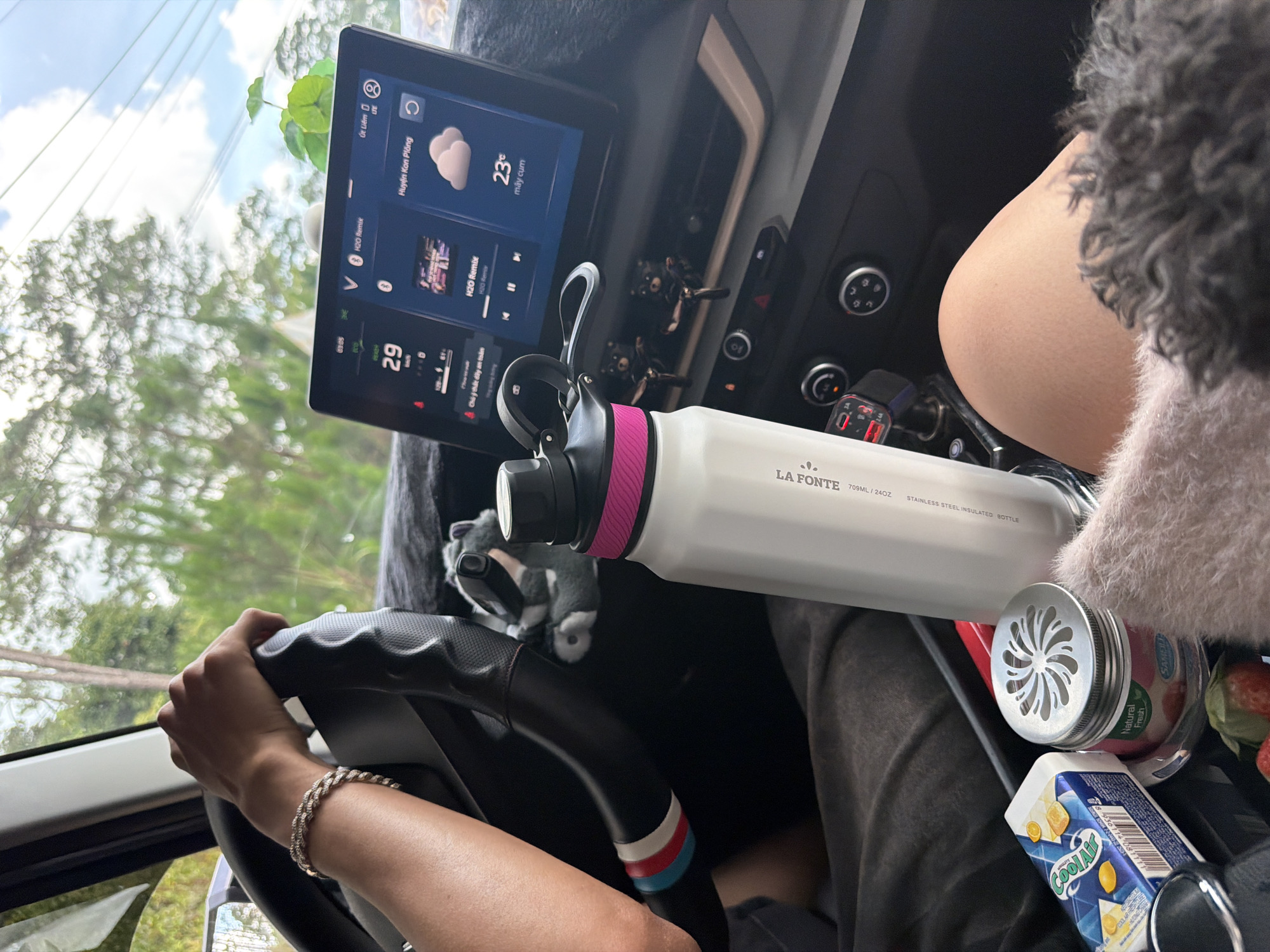 The image captures an inside view of a car, likely in Măng Đen, Vietnam, focusing on the dashboard and parts of the front cabin. A person's hands are visible on the steering wheel, suggesting they are driving. Another person's leg and a furry item (possibly a pet or blanket) are seen on the passenger side.
The central infotainment screen displays several pieces of information: the location "Huyện Kon Plông" (Kon Plông District, where Măng Đen is situated), a weather icon indicating clouds and a temperature of "23° mây cum" (23°C cloudy). Music, "H2O Remix," is shown playing, and a speedometer indicates "29 km/h". A safety message, "Chú ý thức dậy an toàn" (Pay attention, wake up safely), is also visible.
Various objects are arranged around the car's interior. A white "LA FONTE 709ML / 24OZ STAINLESS STEEL INSULATED BOTTLE" with a pink and black cap is prominently placed. Nearby, a small stuffed animal, possibly a badger or raccoon, sits on the dashboard. A USB charger with a red digital display showing "0.0" is plugged in. Snacks are visible on the passenger side, including a carton of "COOLAIR" juice and a container labeled "SAMEL Natural Fresh." Several strawberries are also present. The driver wears a silver-colored bracelet on their right wrist. The setting suggests a daytime journey under partly cloudy skies.