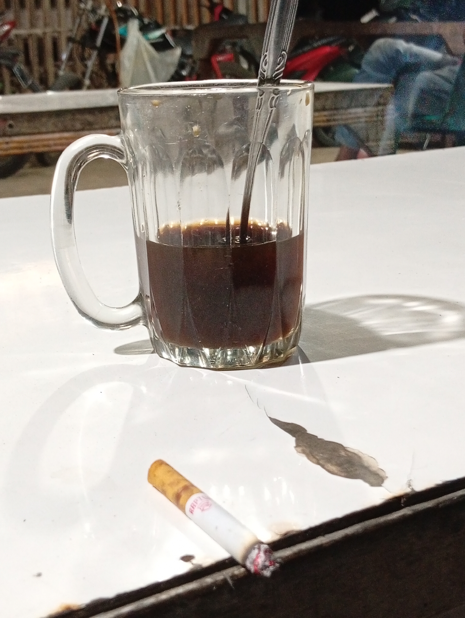 The image displays a close-up, ground-level view of a white table in Lhokseumawe, Indonesia. A clear glass mug, approximately half-filled with a dark brown liquid, occupies the upper-left to mid-right section. A silver spoon, featuring patterned ornamentation on its handle, is immersed in the liquid. To the lower right of the mug, a lit cigarette rests on the table. The cigarette exhibits a yellowish-brown filter, a white body with a partially visible red logo, and an actively burning, ash-covered tip. The white table surface shows notable wear and damage, including cracks, peeling paint, and dark discolored patches, particularly along the bottom edge where it transitions to a dark, possibly wooden, border. Harsh lighting from an unseen source creates distinct shadows on the table, including the mug's shadow with refracted light patterns from its base and the shadow of the cigarette. The background is significantly out of focus, revealing blurred elements such as parts of parked vehicles, possibly motorcycles, a slatted wooden or bamboo structure, and indistinct figures, one of whom appears to be wearing blue jeans.