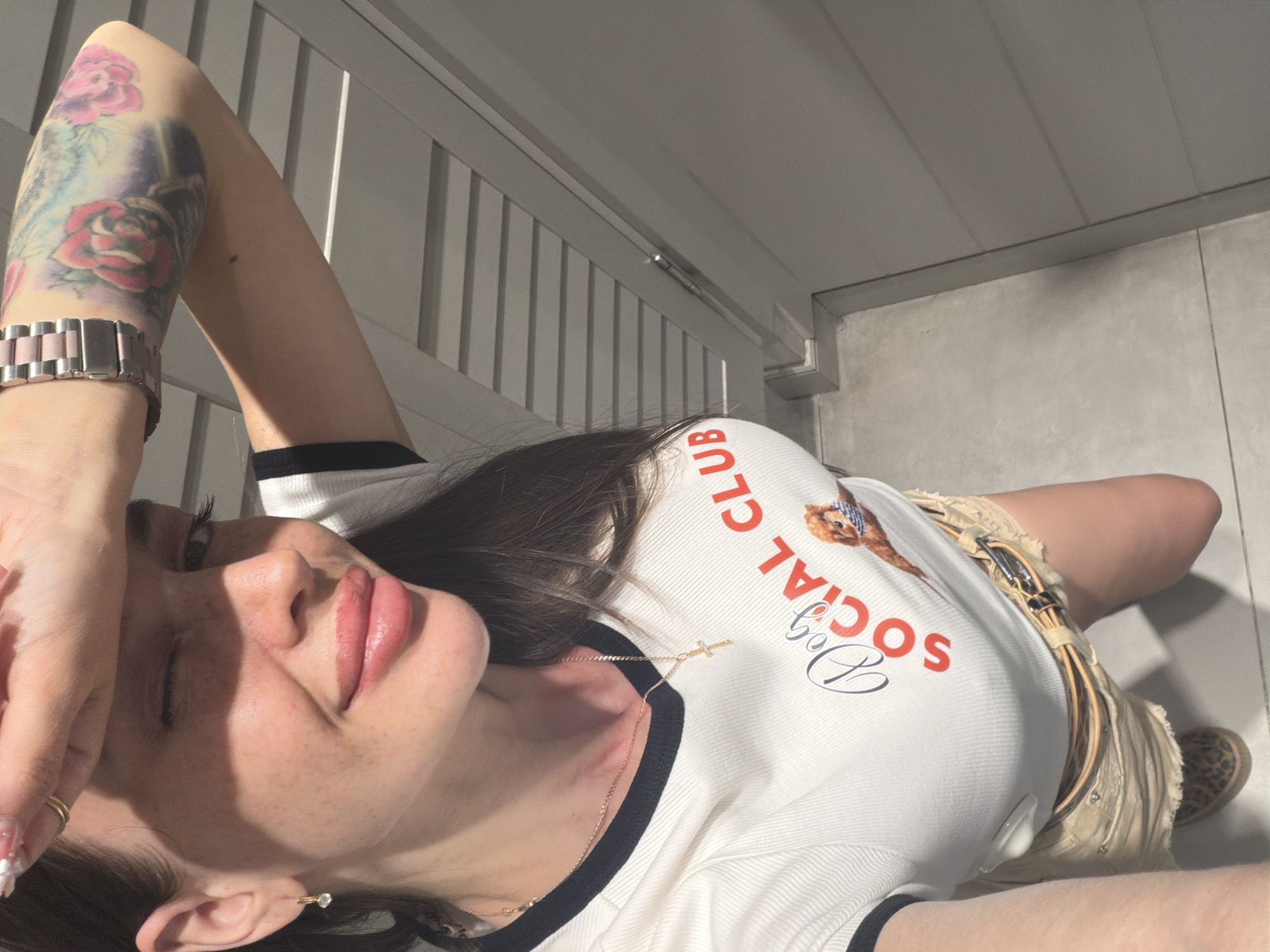 A medium shot shows a woman from the chest up, taking a selfie. She has long dark hair, and is wearing a white ribbed t-shirt with a black trim. The t-shirt has a graphic of a dog and the words "Dog Social Club" printed on it. She is also wearing light-colored shorts and a leopard-print shoe is visible. She has a gold cross necklace on, and her right arm is raised, covering part of her face. A tattoo of roses is visible on her arm, and she is wearing a watch. The background appears to be an indoor setting with a wall that has horizontal lines. The flooring is tiled.