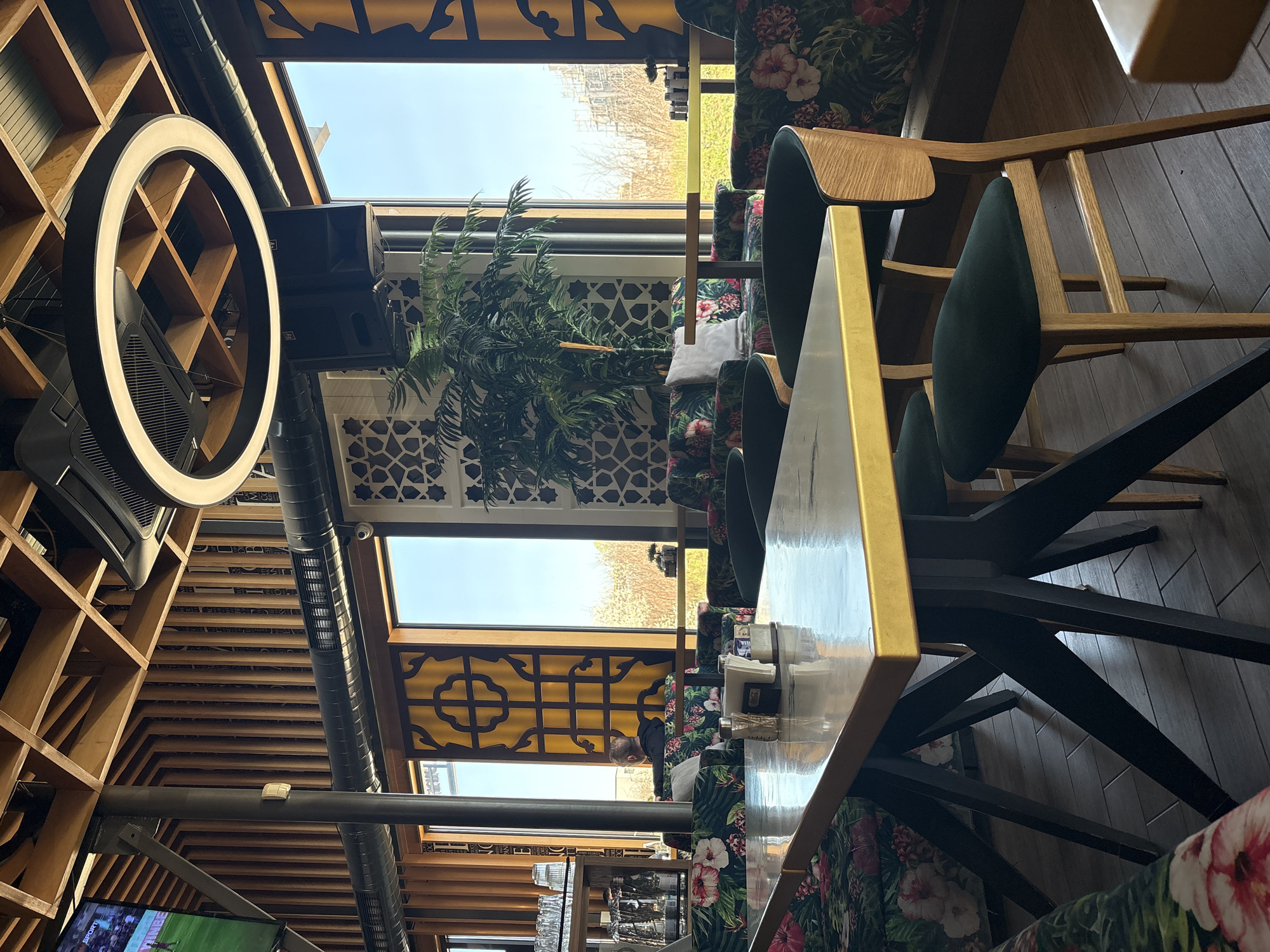 The image shows the interior of a restaurant or café in Sofia, Bulgaria. The interior design features a modern aesthetic with wooden latticework on the walls and ceiling, a ring light fixture, and a long table with gold trim. The chairs have dark green seats and floral upholstery on the backrests. A potted plant is visible near a window. A person is sitting at the table in the distance, appearing to be looking at something. A TV screen can be seen in the lower left corner of the frame. The setting is an enclosed indoor space with natural light filtering through the windows.