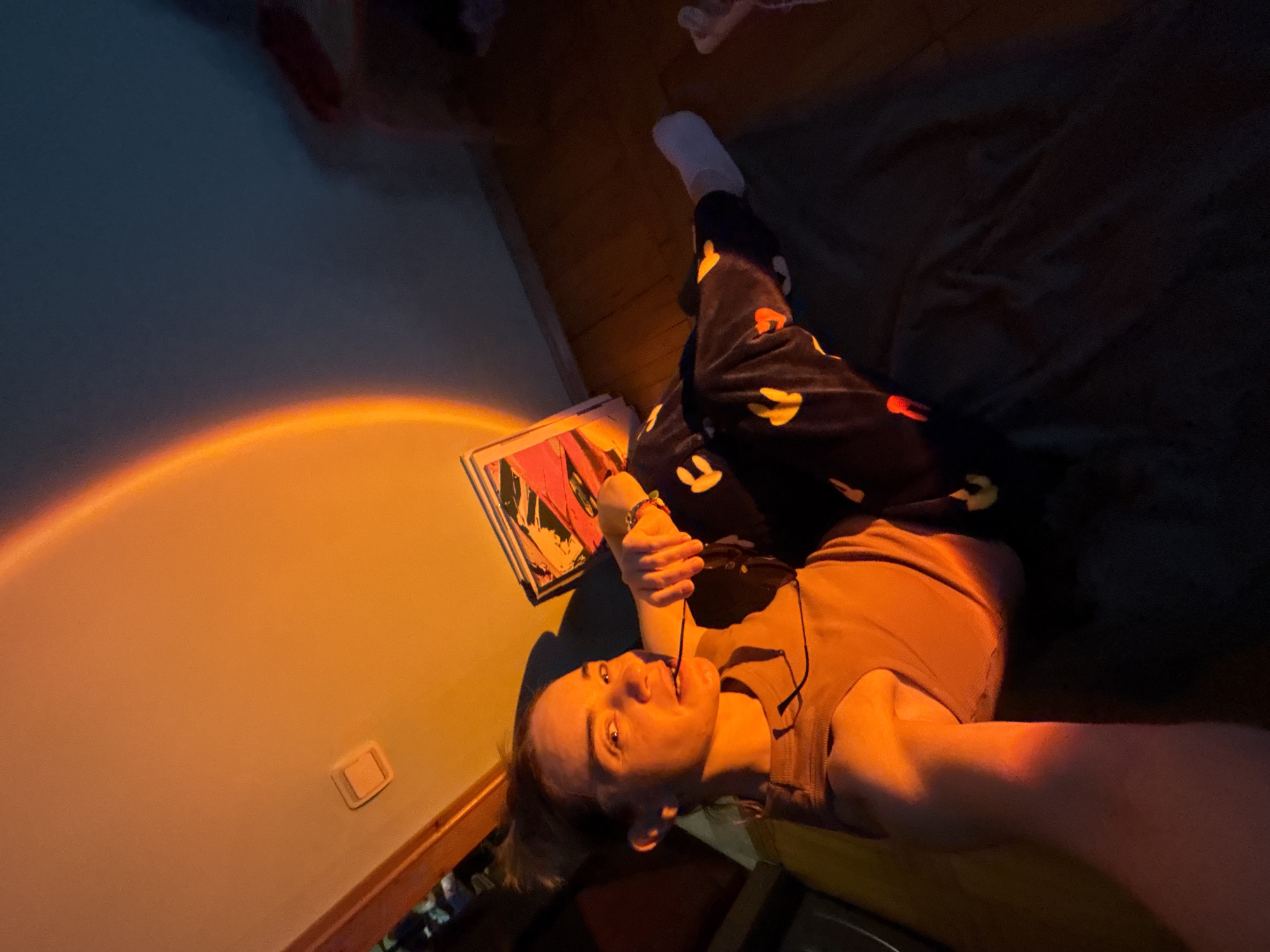 A person lies on their back on a bed or similar surface, holding glasses in their mouth, in an indoor setting. The person is wearing patterned pants and a tan tank top. A white sock is visible on one foot. An orange light source illuminates part of the wall and floor, casting an arc. There is a framed print or artwork near the person. The room appears to be dimly lit aside from the orange light. The location is unknown.
