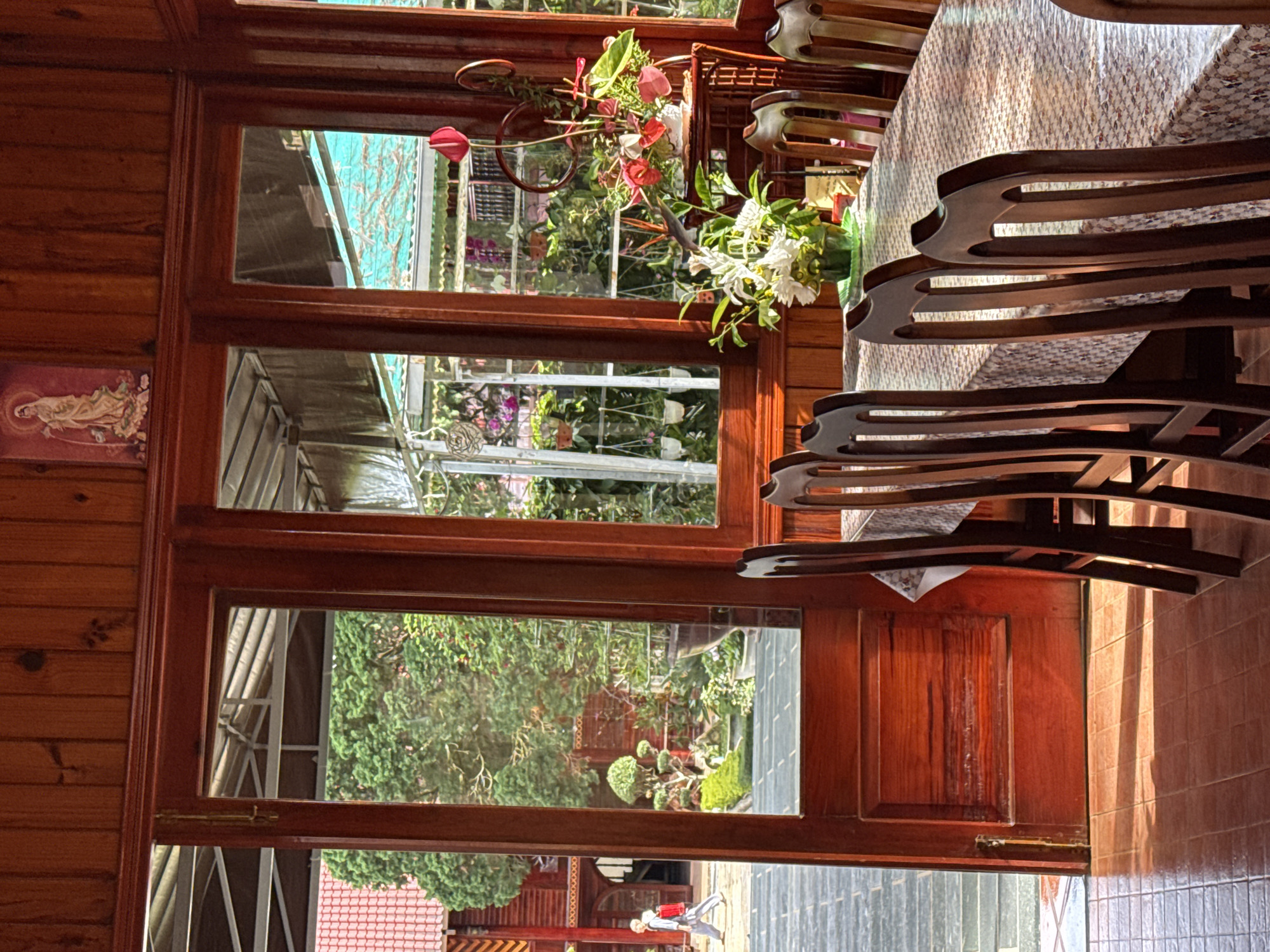 A wooden-paneled room with a large glass door and window offers a view of a garden. A long dining table covered with a patterned tablecloth is set with chairs. Sunlight streams in, creating shadows on the tiled floor. In the garden, there are trees, potted plants, and architectural structures. A person in a light-colored robe and a red backpack walks through the garden. The room appears to be in Da Lat, Vietnam, suggested by the architectural style and the presence of what seems to be a Buddhist deity depicted in an image on the wall.