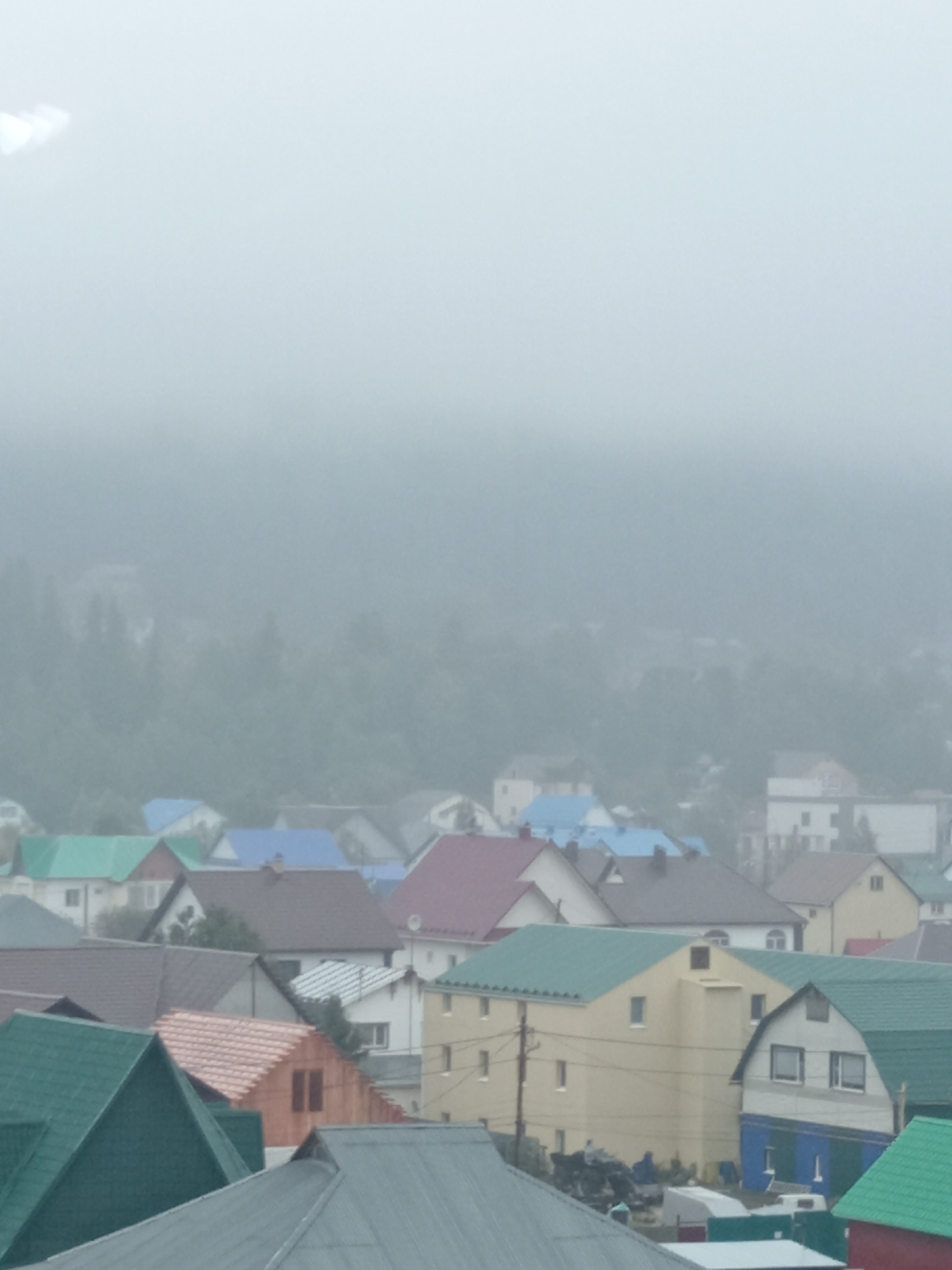 The image depicts a residential area with numerous houses, most of which have brightly colored roofs in shades of green, blue, red, and brown. The houses are of varying architectural styles and sizes, suggesting a mix of older and newer constructions. Some houses appear to be two stories tall, while others are single-story dwellings. The foreground is dominated by the roofs of several houses, with one large, dark gray metal roof in the bottom center of the frame. Behind these, a cluster of houses with vibrant green and red roofs are visible. Further back, more houses with blue and brown roofs are interspersed with buildings that are less distinct due to the foggy conditions. In the background, a dense forest covers a hillside, and the entire scene is shrouded in a thick fog or mist, obscuring details of the landscape and distant structures. The overall impression is of a somewhat rural or suburban setting on an overcast or foggy day. No people are clearly visible in the image. The location is identified as Khanty-Mansiysk, Russia.