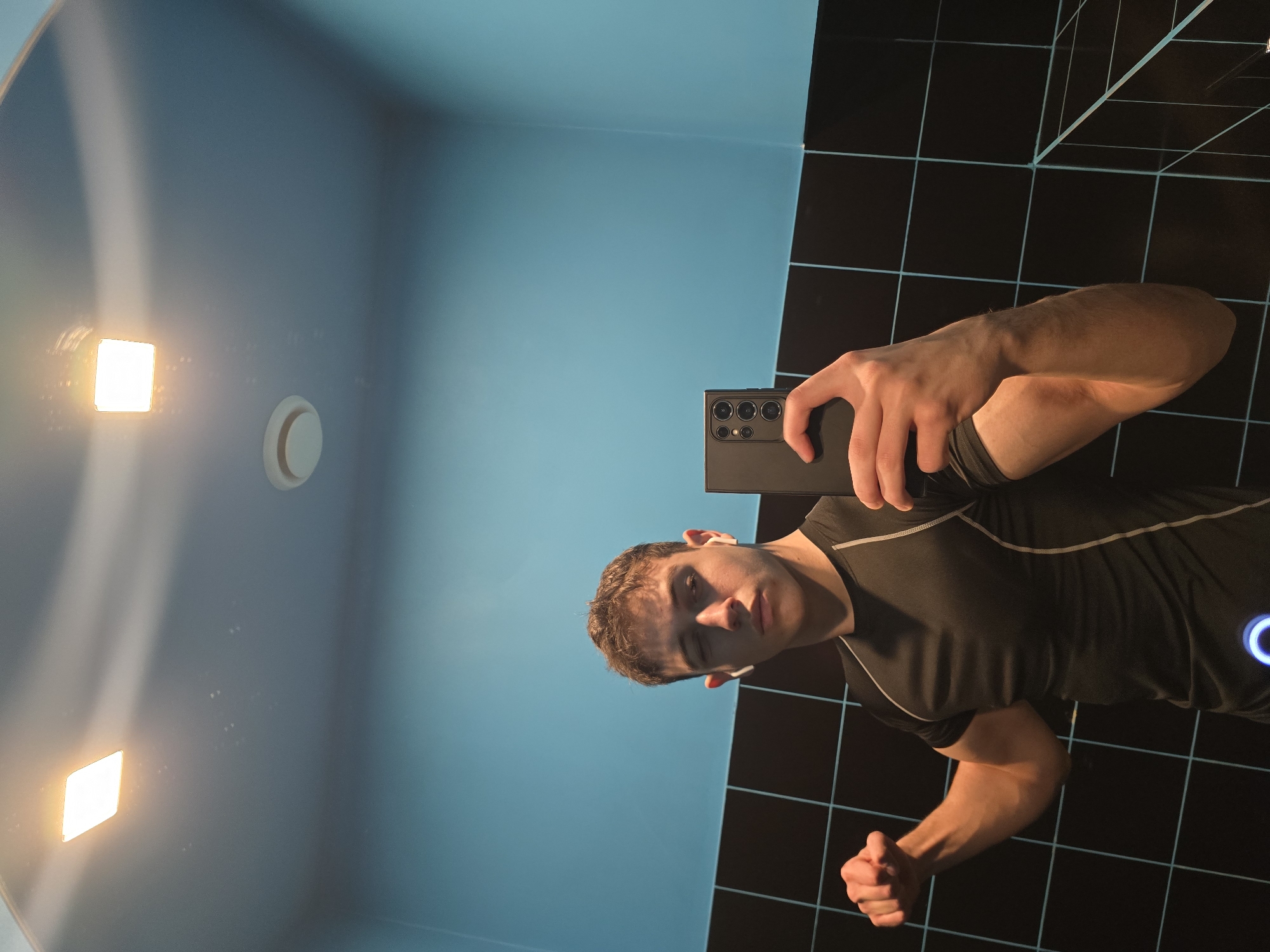 A young adult male is depicted in a mirror selfie taken within a restroom or changing facility in Yerevan, Armenia. The subject, with short brown hair and wearing a black athletic t-shirt with light-colored piping, flexes his right bicep while his left hand holds a dark-colored smartphone, identified by its vertical triple-lens camera array, to capture the image. White wireless earbuds are visible in both of the subject's ears. The reflected environment features light blue upper walls and ceiling, with the lower walls covered in black square tiles separated by light blue grout lines. Two square, recessed ceiling lights emit warm illumination, and a circular white ventilation fan is visible on the upper wall. The subject maintains a neutral facial expression, with his left eye appearing partially closed.