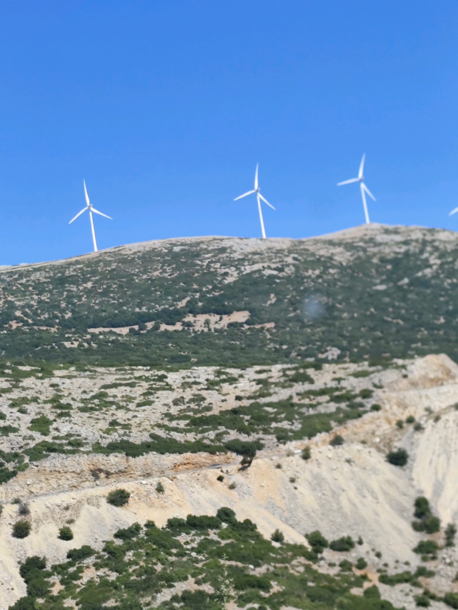 The image shows a clear blue sky above a barren, rocky hillside in the Municipal Unit of Achladokampos, Greece. Several white wind turbines with three blades each are visible atop the hill, silhouetted against the sky. The hillside is covered with sparse green vegetation, with a winding dirt path visible in the foreground. The terrain appears rugged and arid, typical of a Mediterranean landscape.