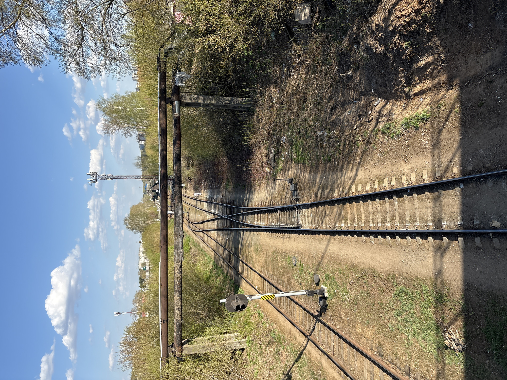 The image shows a railway track in the city of Chaikovsky, Russia. The track is viewed from an elevated position, likely a bridge or overpass. The scene includes:
* **Railway Tracks:** Two parallel tracks that converge at a switch point further down the line.
* **Vegetation:** Trees and bushes lining both sides of the track, with signs of early spring foliage.
* **Infrastructure:** An overhead structure spanning the tracks, possibly carrying utilities or serving as part of the railway infrastructure.
* **Signaling:** A railway signal post with a horizontal arm displaying a combination of black, yellow, and white stripes.
* **Sky:** A blue sky with scattered white clouds.
* **Ground:** A dirt and gravel bed surrounding the tracks.
* **Shadows:** Shadows cast by the overhead structure and nearby trees.
There are no people visible in the image. The scene depicts a functional, if somewhat overgrown, railway environment.