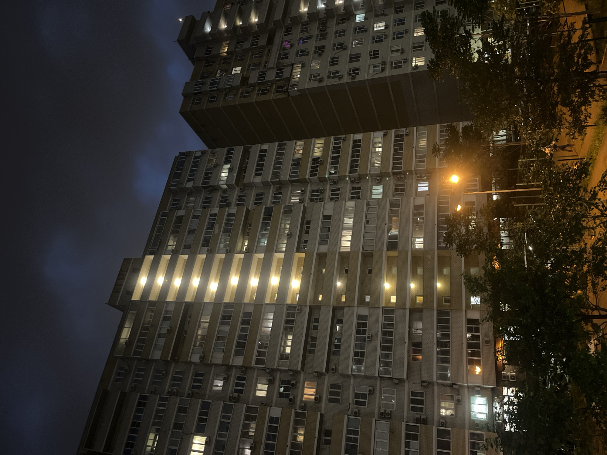 The image shows a nighttime view of a residential building in Krasnodar, Russia. The building is tall and multi-storied, featuring a repetitive pattern of windows, many of which are illuminated. Air conditioning units are visible on the exterior walls. A streetlight illuminates the right side of the building and the adjacent trees, while the sky above is dark and cloudy.