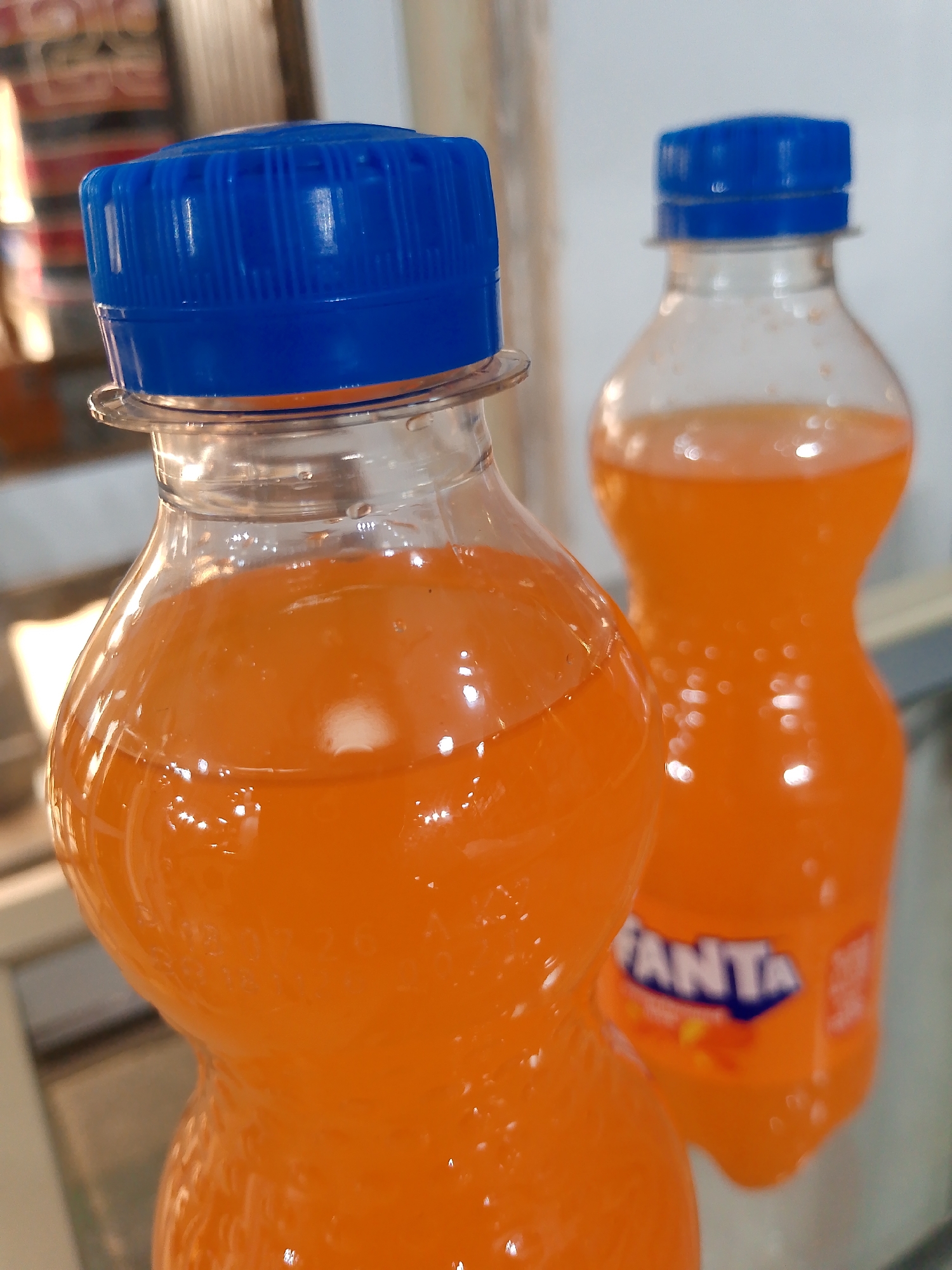The image shows two plastic bottles filled with an orange-colored beverage. The bottle in the foreground is closer to the camera and is larger, while the bottle in the background is smaller and slightly out of focus. Both bottles have blue plastic caps. The background is blurred, but appears to be indoors with some shelving or furniture visible. The scene is brightly lit, with highlights reflecting off the liquid in the bottles. The location is identified as Bukuru, Nigeria.
