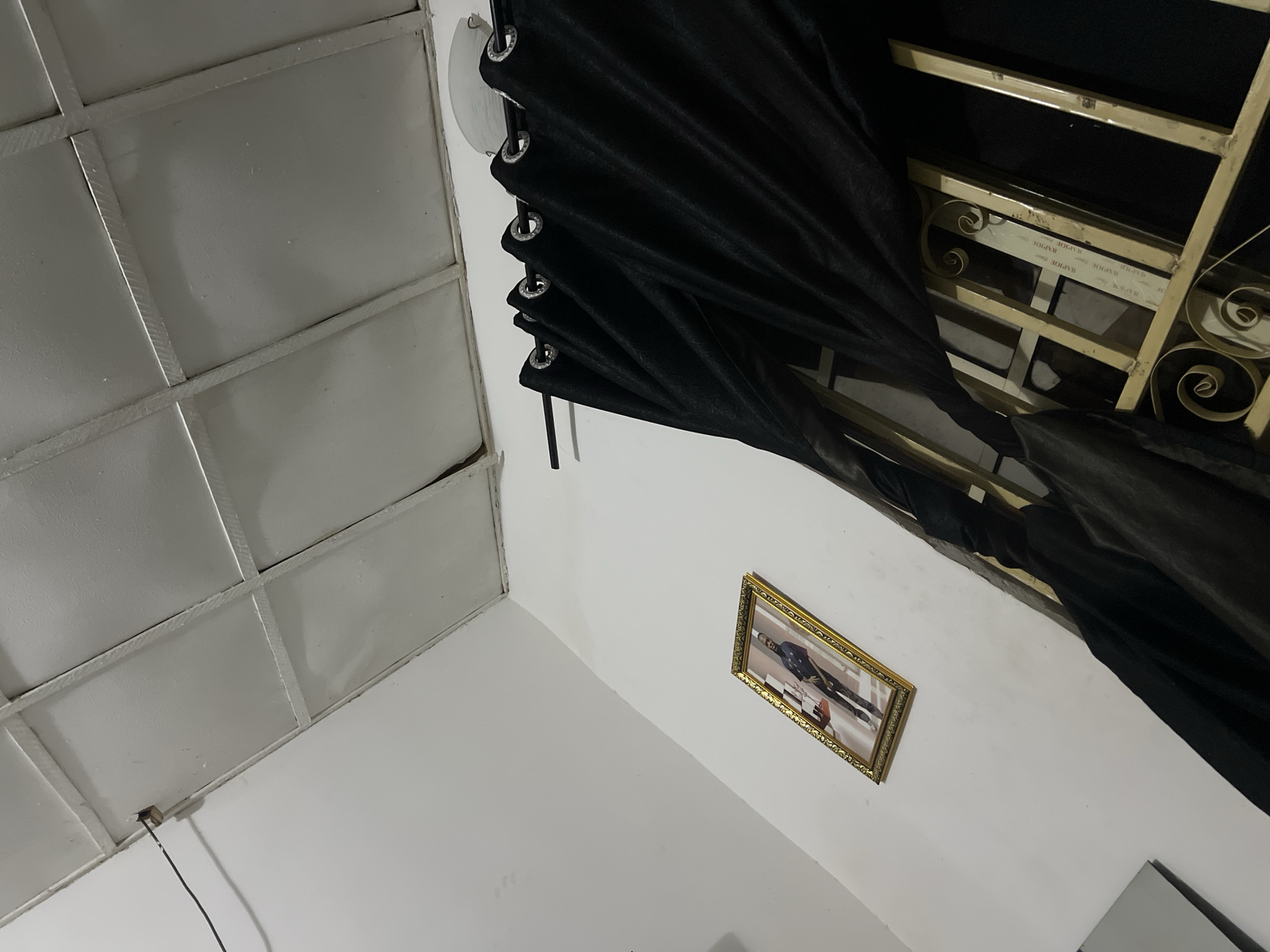 The image depicts an interior scene, likely a room in Howayi, Nigeria. The perspective is upward, showing a white, paneled ceiling with a visible electrical wire and an outlet. On the white wall below, a framed photograph of a man in formal attire is hung. To the right, black curtains with grommets are drawn across a window that has a decorative metal grille. The overall impression is of a domestic or office space.