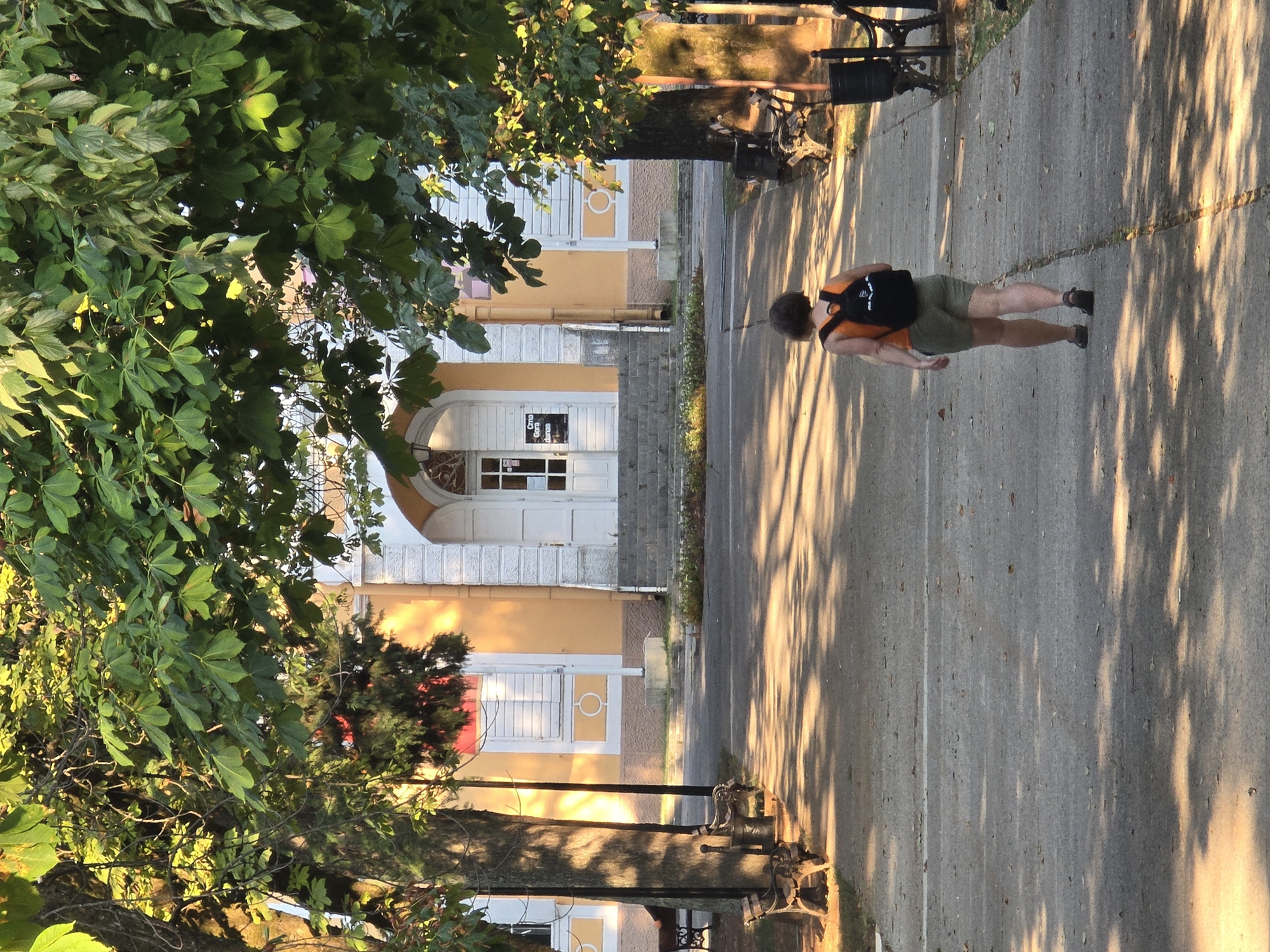 A woman with short, brown hair walks away from the camera down a sun-dappled paved pathway. She is wearing an orange tank top, olive green shorts, and dark sandals. A black backpack with a white logo is on her back. The pathway is lined with benches on either side. To the left, a building with an orange facade, white trim, and closed shutters is visible behind trees. To the right, more trees and benches are present, with a trash can visible next to one bench. The building has a white portico with stairs leading up to a white double door. The scene appears to be in the Kruševac Park in Podgorica, Montenegro, as indicated by the distinctive architecture of the building which resembles the former Kruševac Cinema or a similar historical structure within the park. The time of day is likely late afternoon or early evening, due to the long shadows cast by the trees.