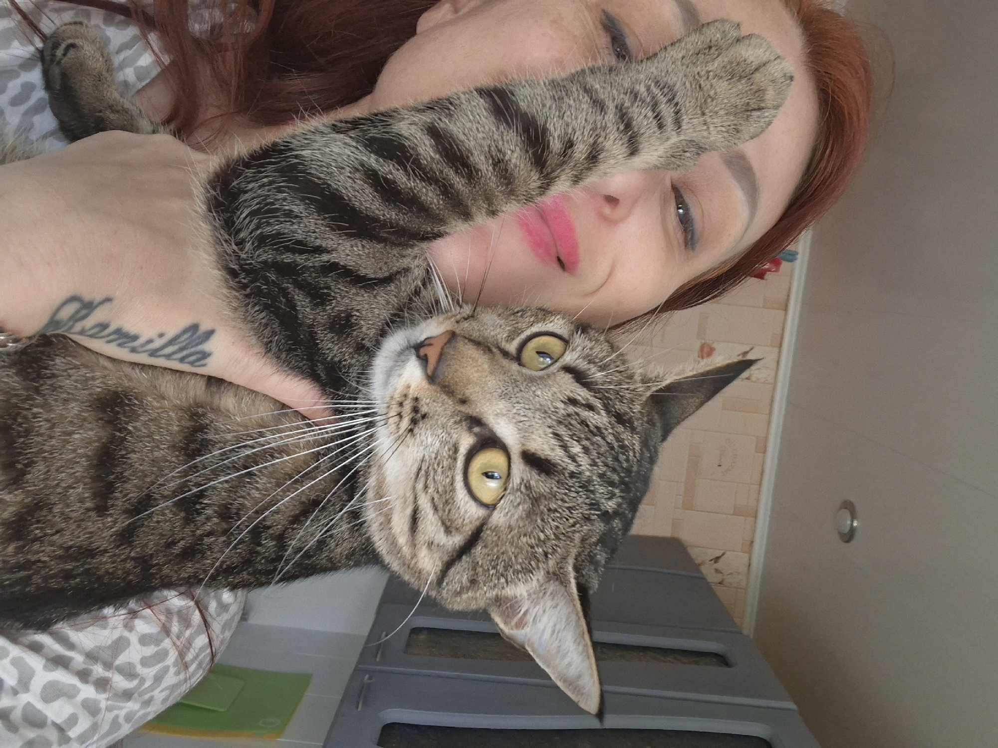 A medium shot shows a person holding a tabby cat in what appears to be a kitchen. The person, with reddish-brown hair, is looking towards the camera with a smile and pink lipstick. Their left arm is raised, partially obscuring their face, with the cat's paw resting on their forehead. The cat is looking to the left of the frame, its yellow eyes wide. The background features kitchen cabinets and patterned wallpaper. A tattoo of the word "Camilla" is visible on the person's forearm.