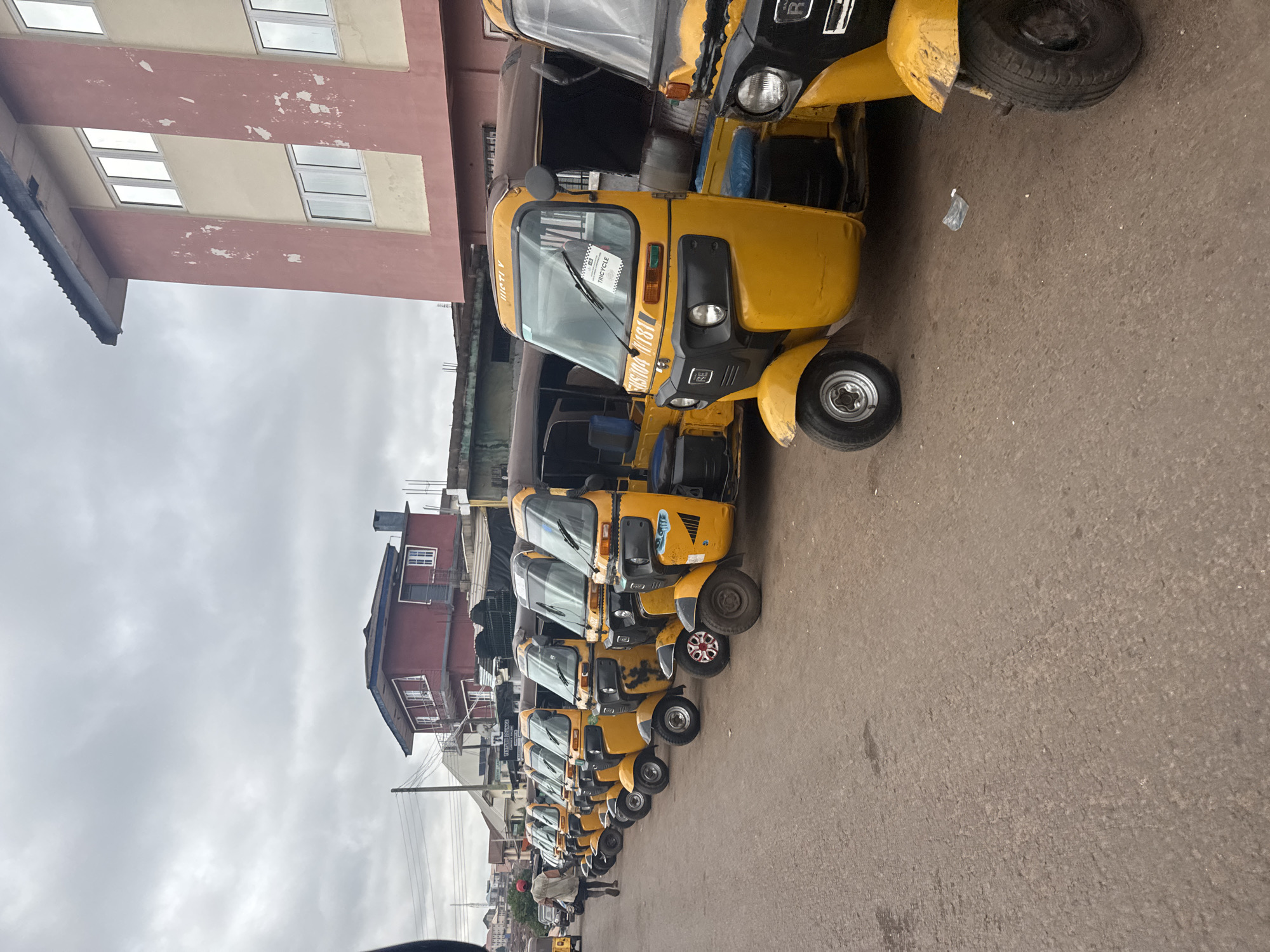 The image depicts a row of yellow tricycles, commonly known as "Keke Napep" or "Okada," parked on a paved street in Enugu, Nigeria. The tricycles are arranged in a line, facing towards the right side of the frame. The foreground shows the front wheels and lower bodies of several tricycles, with the ground appearing to be a mixture of asphalt and pavers.
In the background, there are buildings, including a prominent red and beige building on the right with multiple windows. To the left, there are other structures, some appearing to be shops or commercial establishments, with awnings and signs visible. Power lines and poles are also present in the background, indicating an urban setting.
One man, wearing a red cap and a light-colored shirt, is walking away from the tricycles towards the left side of the image. The sky is overcast with clouds, suggesting a potentially rainy or dull day. The overall scene appears to be a commercial area where these tricycles are likely used for transportation services. The specific location within Enugu is not explicitly identified in the image, but it is a street scene with parked vehicles and buildings.