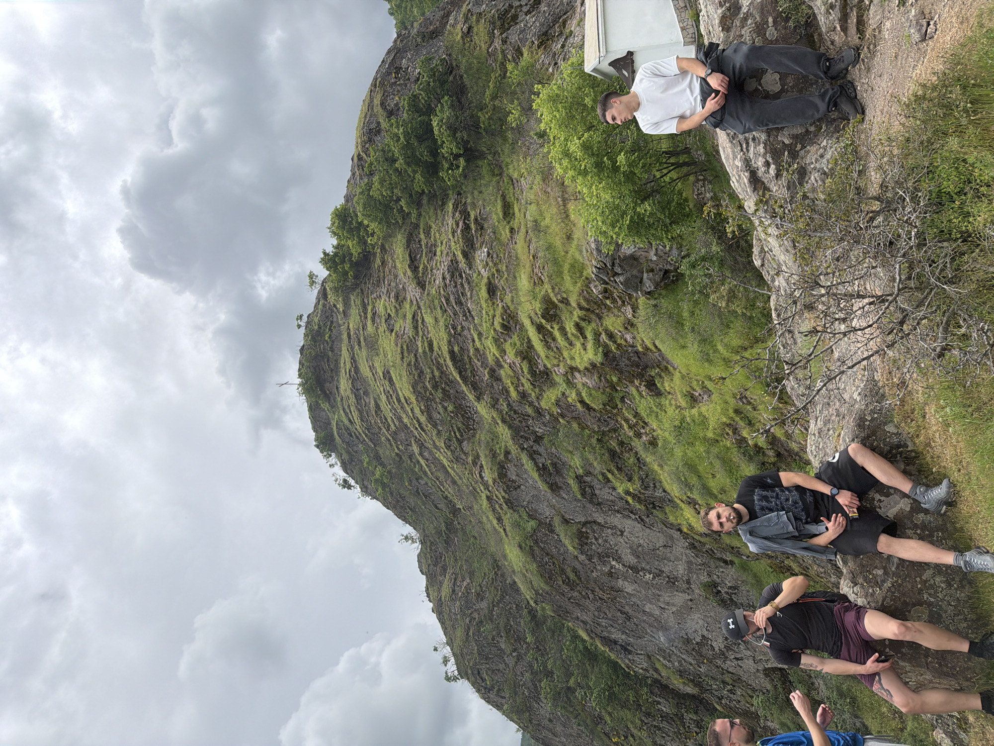 The image shows a group of four people situated on a rocky terrain near a mountain in Ustina, Bulgaria. The sky is overcast with cloudy weather.
One man in a white t-shirt and dark pants sits on a ledge, turned away from the camera. Two men are lying down on rocks in the foreground. One is wearing a black t-shirt and shorts, holding a gray jacket and looking at the camera. The other man in a black t-shirt, shorts, and a baseball cap is below him. The fourth person is partially visible at the bottom, wearing a blue jacket.
The background features a large, steep mountain covered with grass and trees. A small white structure is visible near the top of the cliff.