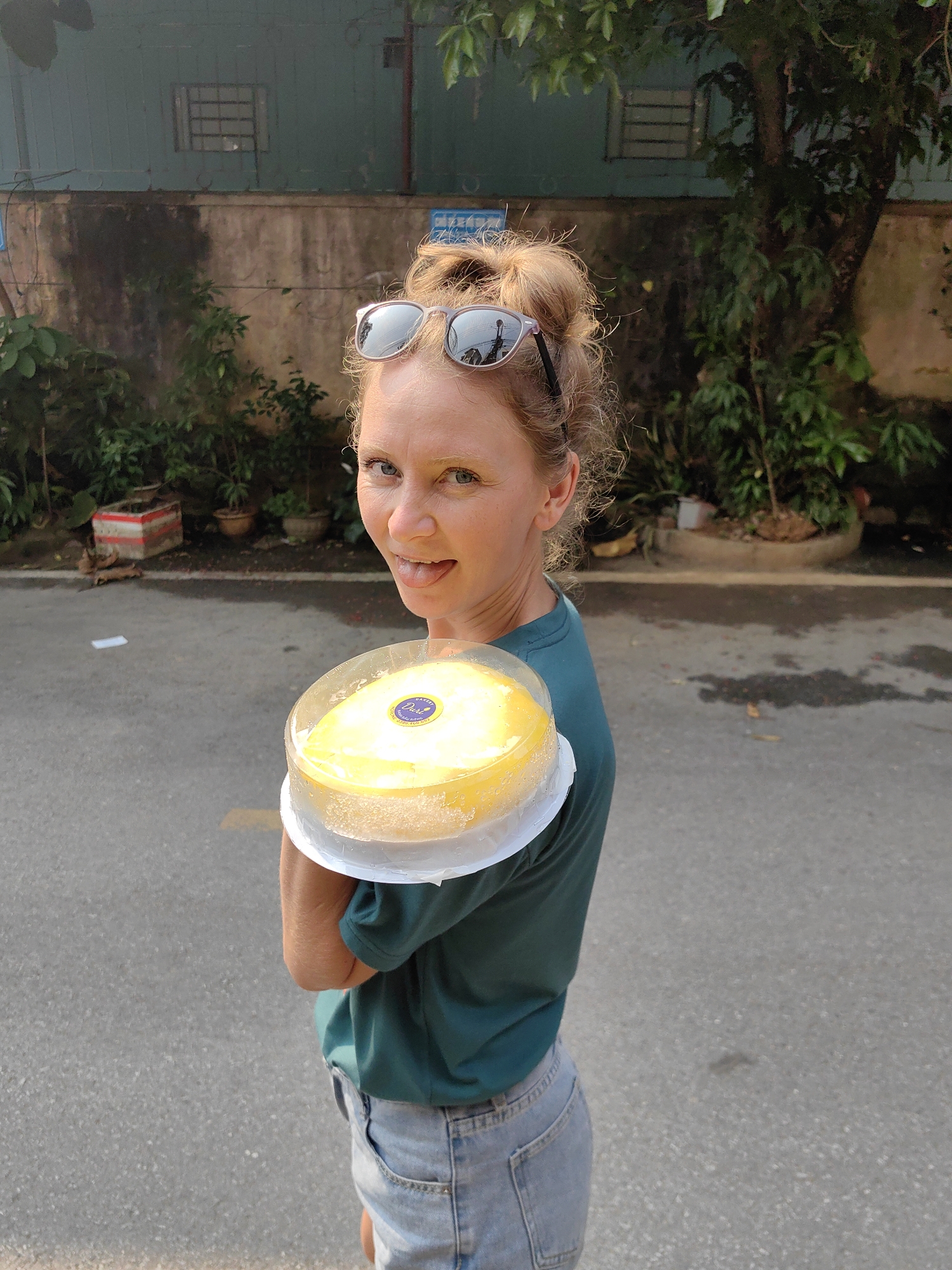 A woman is standing outdoors, holding a cake. She is fair-skinned, with blonde hair pulled up in a messy bun, and is wearing sunglasses on top of her head. She is wearing a dark green t-shirt and light blue jeans. She has her tongue sticking out slightly. The cake she is holding is round, yellow, and in a clear plastic container with a white base. The cake has a blue label with white text on it. She is standing on a gray asphalt street. Behind her is a low building with a faded green wall, with a few windows. In front of the building are various potted plants and trees. Based on prior knowledge, the city of Thành phố Hà Giang, Vietnam, is likely in the background.