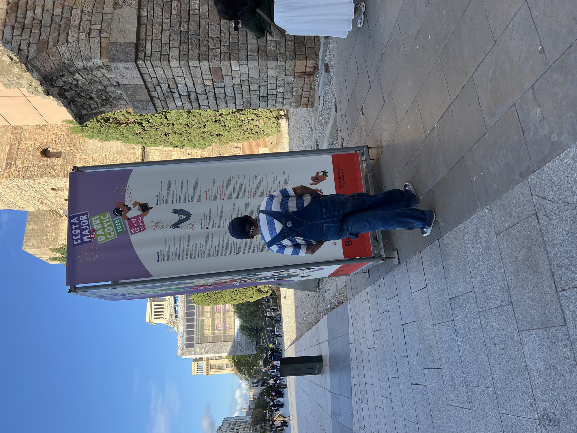 The image captures a street scene in Barcelona, Spain, showcasing an outdoor informational poster advertising the “Festa Major del Barri Gotic 2026” which took place from May 13-17. A man, wearing a striped shirt and blue overalls, is standing in front of the poster, seemingly reading it. The poster is printed with the city of Barcelona logo, "B" and "Fem la Barcelona de la teva vida."
The environment includes a stone wall that might be part of an older building, adjacent to greenery. The pathway, made of gray stone tiles, stretches towards the distance, where we see a green bin and several people walking along the street and buildings in the background. The sky is clear and blue.