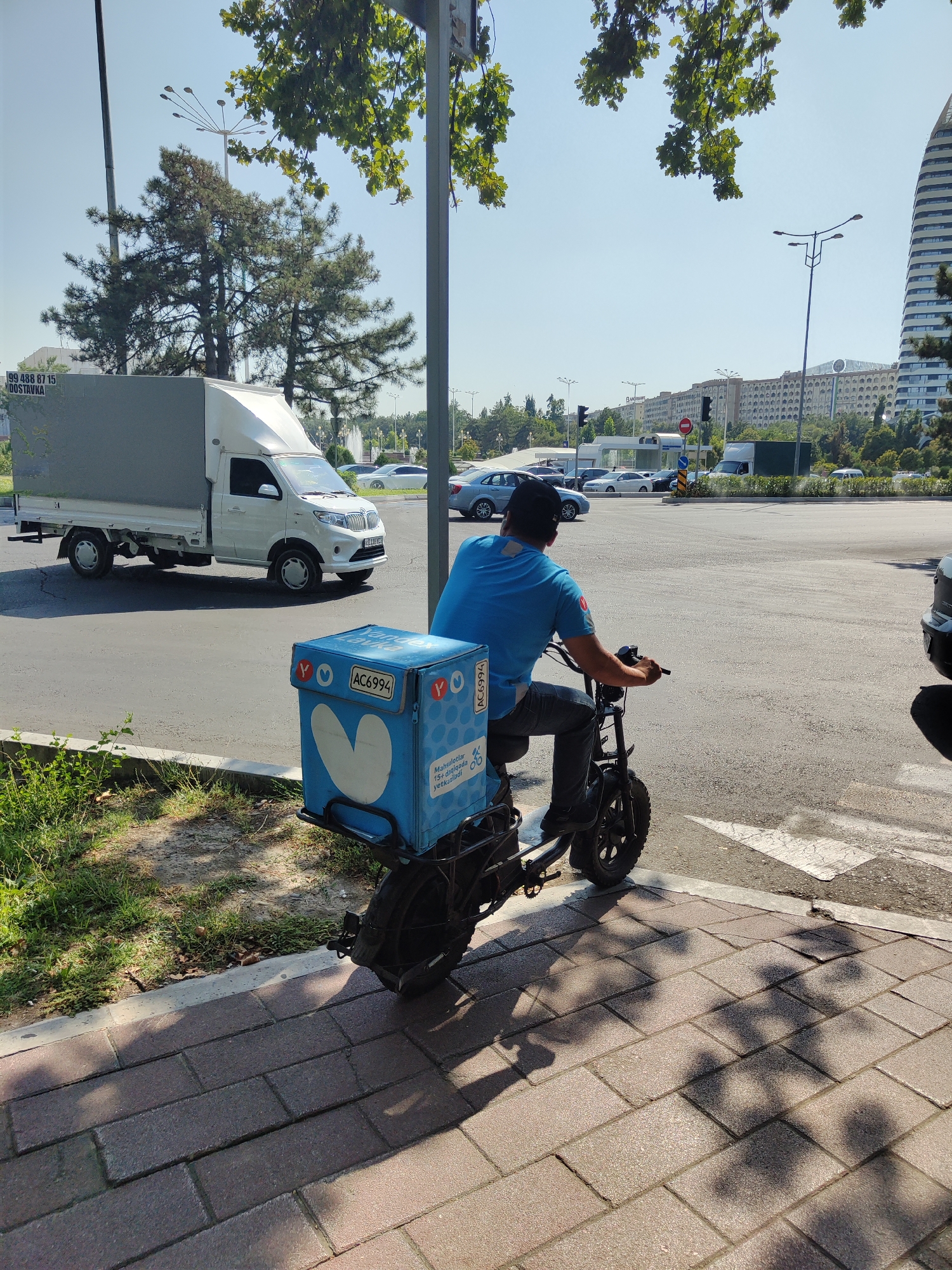 A delivery person on a black electric scooter with a blue insulated delivery box on the back is stopped at an intersection in Tashkent, Uzbekistan. The delivery box has a heart logo and text in Cyrillic. The person is wearing a blue t-shirt and dark pants, with a black cap. In the background, a white truck with a gray cargo box is driving on the road, and other cars are visible at the intersection. A tall, modern building with a curved facade stands on the right side of the frame, and a row of apartment buildings stretches across the middle ground. Trees and streetlights are also present, and the sky is clear and blue. The scene is illuminated by bright sunlight, casting shadows on the sidewalk.