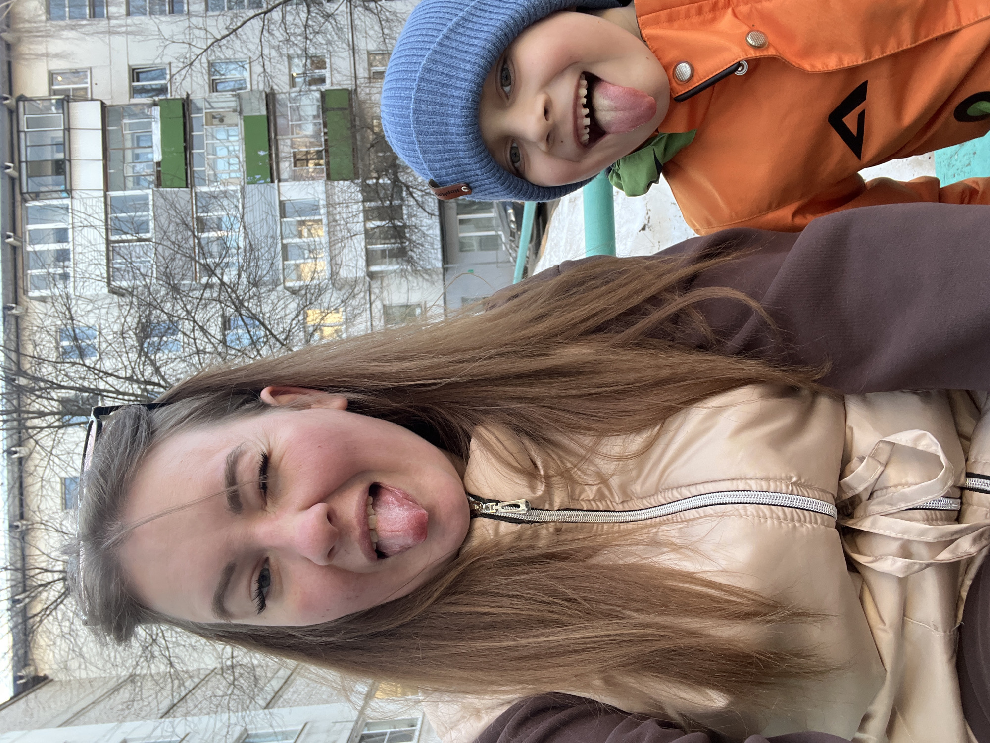 Two individuals, a young boy and a woman, are in the foreground, facing the camera and sticking their tongues out. The boy is wearing a blue beanie and an orange jacket with a green collar. The woman has long brown hair and is wearing a beige jacket and brown sweater.
Behind them, a multi-story residential building is visible. The building is gray. Bare tree branches are visible in front of the building. Based on the architecture and style of the building, the location appears to be in a city in Russia. Given the prompt, the city is Chaikovsky, Russia. The photo was taken outdoors during the day.