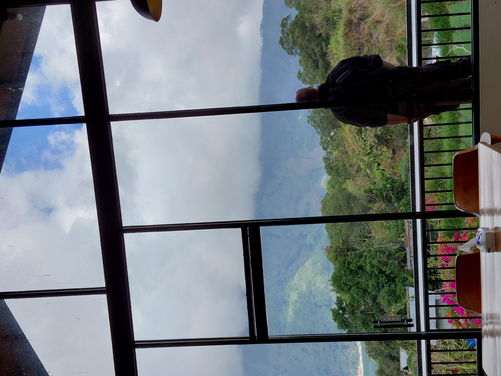 A person stands at a large window with a view of a lush green mountain landscape, likely in Kintamani, Indonesia. The window is divided into panes by dark frames, and the view outside includes a body of water in the distance, surrounded by trees and a misty mountain range. In the foreground, a wooden table is visible with a tissue box on it, and two wooden chairs are partially seen. The person is facing away from the camera, looking out at the scenery, wearing dark clothing and a backpack. The sky is partly cloudy.