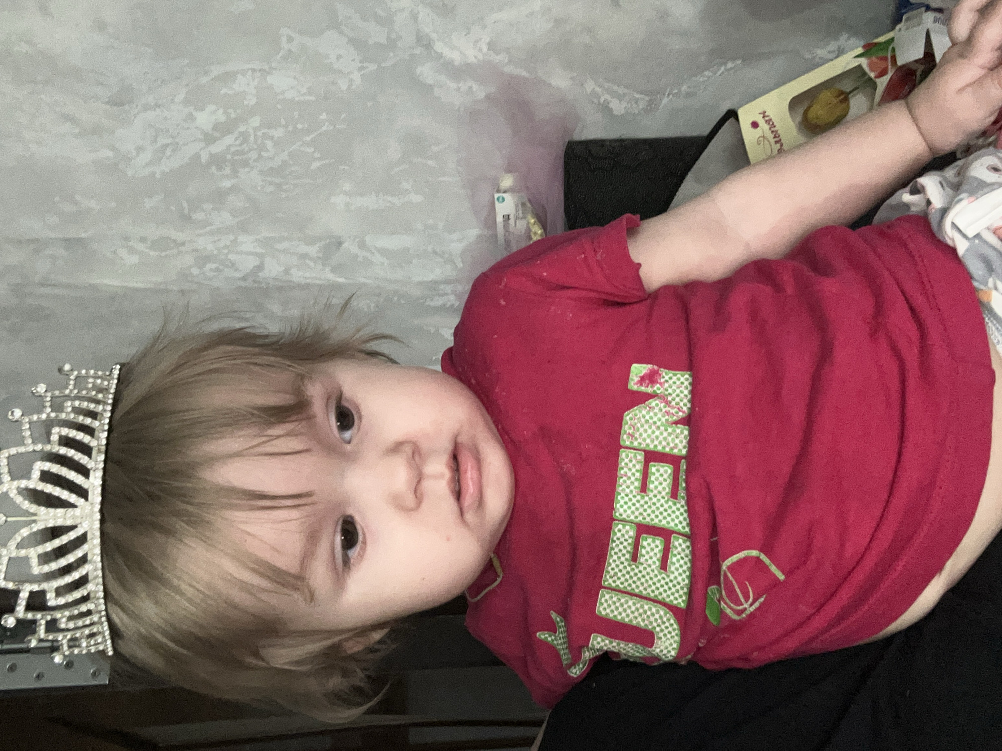 A young child is lying down wearing a red t-shirt with the word "QUEEN" printed on it in green lettering. A silver tiara is on the child's head. The background includes a patterned gray wall and a partially visible box with images of fruit on it. The location is possibly in or around the city of Ольховка, Russia.