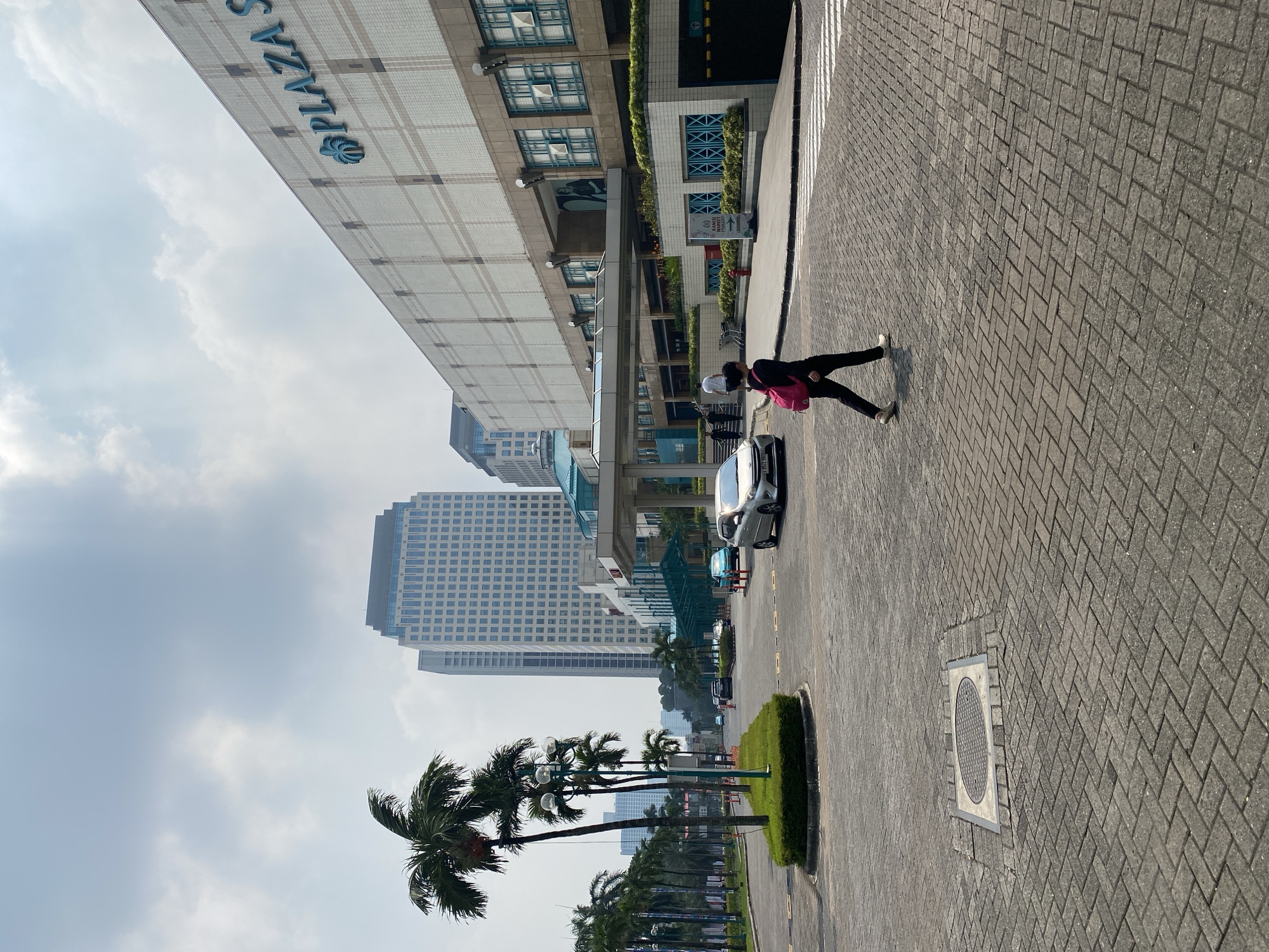 This is a daytime, eye-level, outdoor, street-level photograph taken in Central Jakarta, Indonesia, on a paved street. In the foreground, a person wearing black pants, a black shirt, and a pink backpack walks from left to right across a crosswalk. To the right is a large, modern building with a beige facade, featuring a blue "PLAZA" logo and decorative tile patterns. The building has multiple windows with light blue frames and a covered entrance. A silver sedan is parked on the right side of the street. In the middle ground, several cars are driving on a multi-lane road lined with palm trees. To the left, a circular green lawn with a light post is situated in the middle of the road. In the background, tall, modern office buildings with grid-like windows dominate the skyline, with a cloudy, bright sky above.